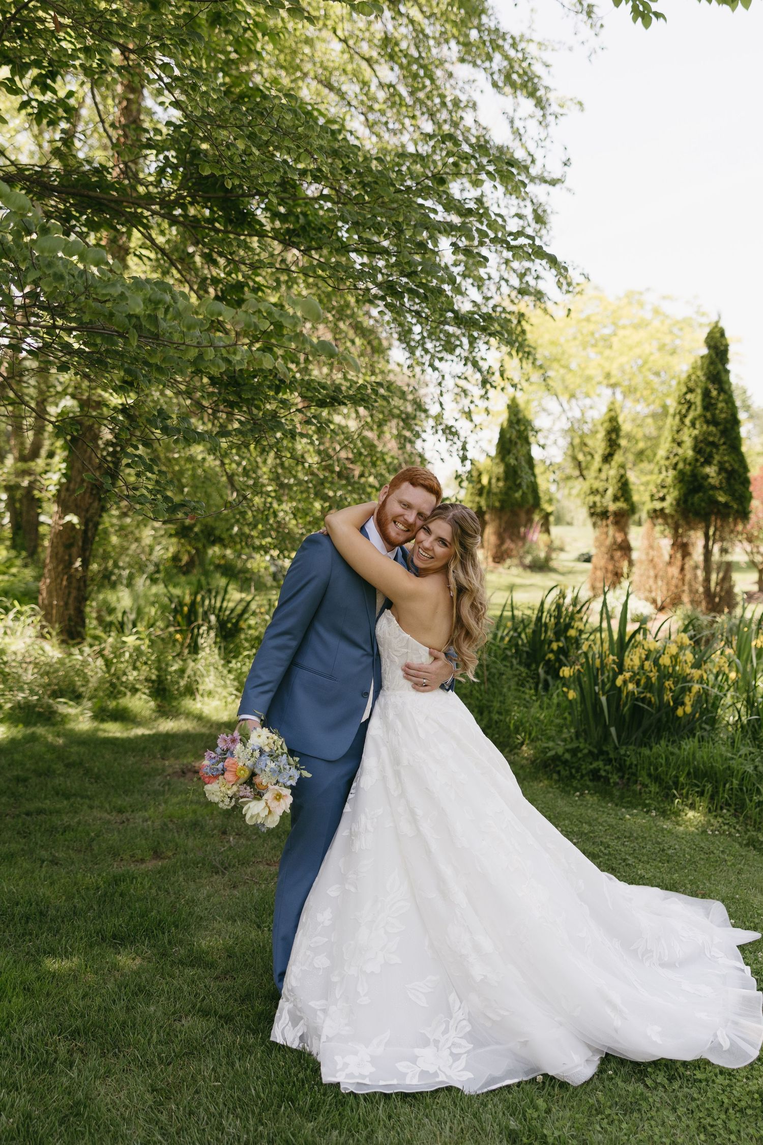 A bride and groom embrace in a lush garden, she holds a colorful bouquet in a white gown, he wears a blue suit.