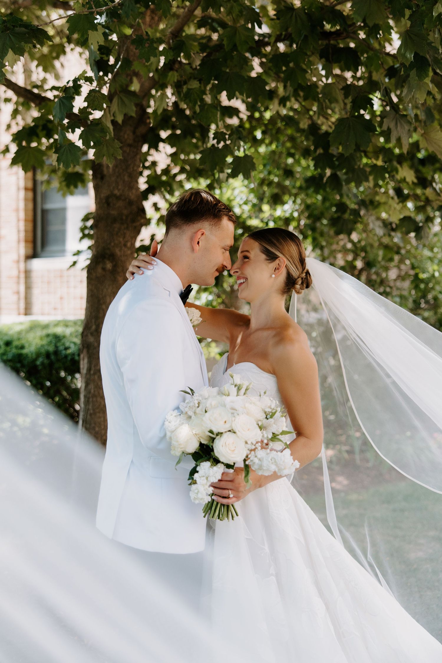 Bride and groom share a romantic moment under a tree, veil flowing in the breeze on wedding day.