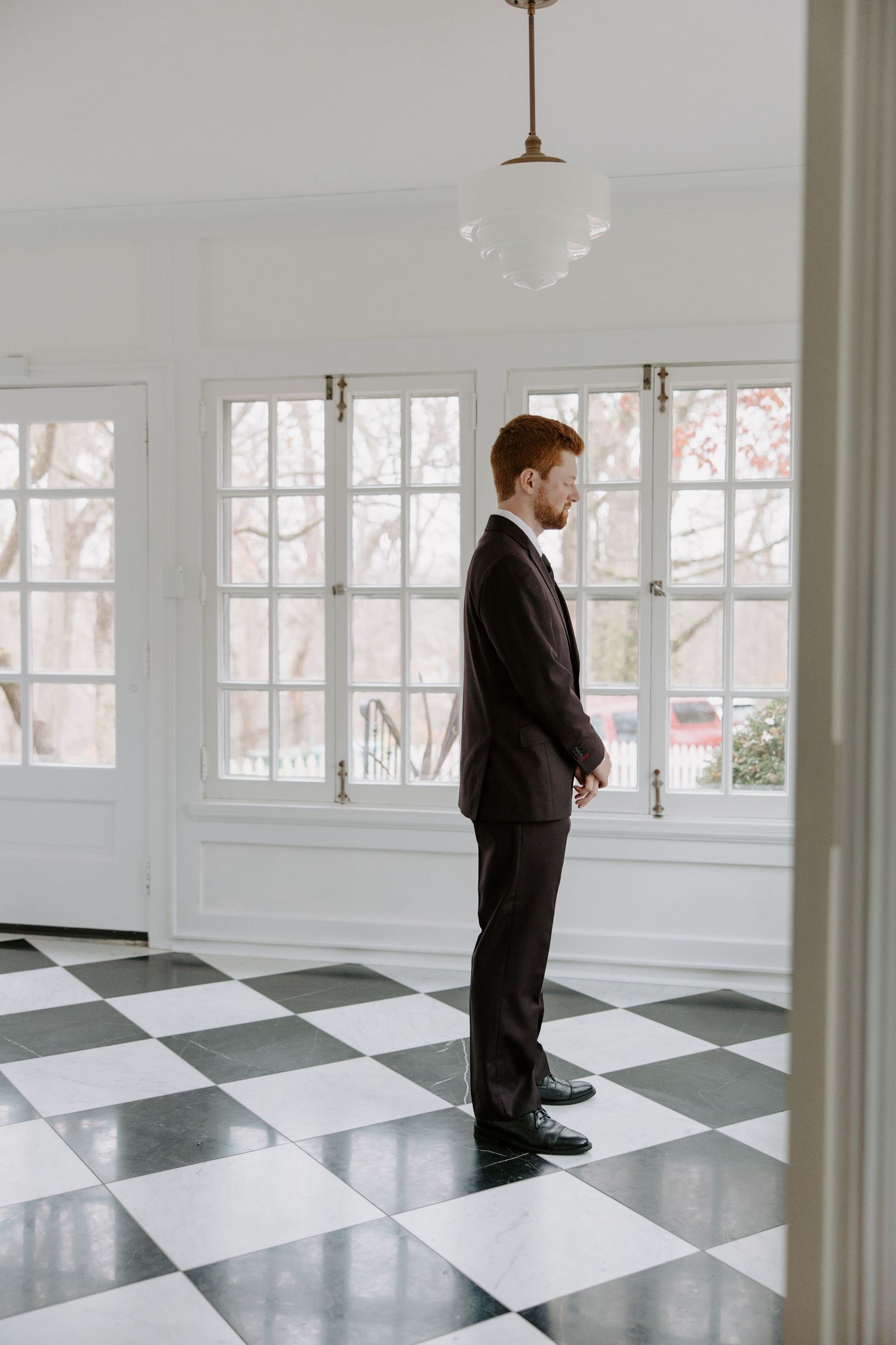 Groom in dark suit standing on black and white checkered floor in bright white room before wedding ceremony.