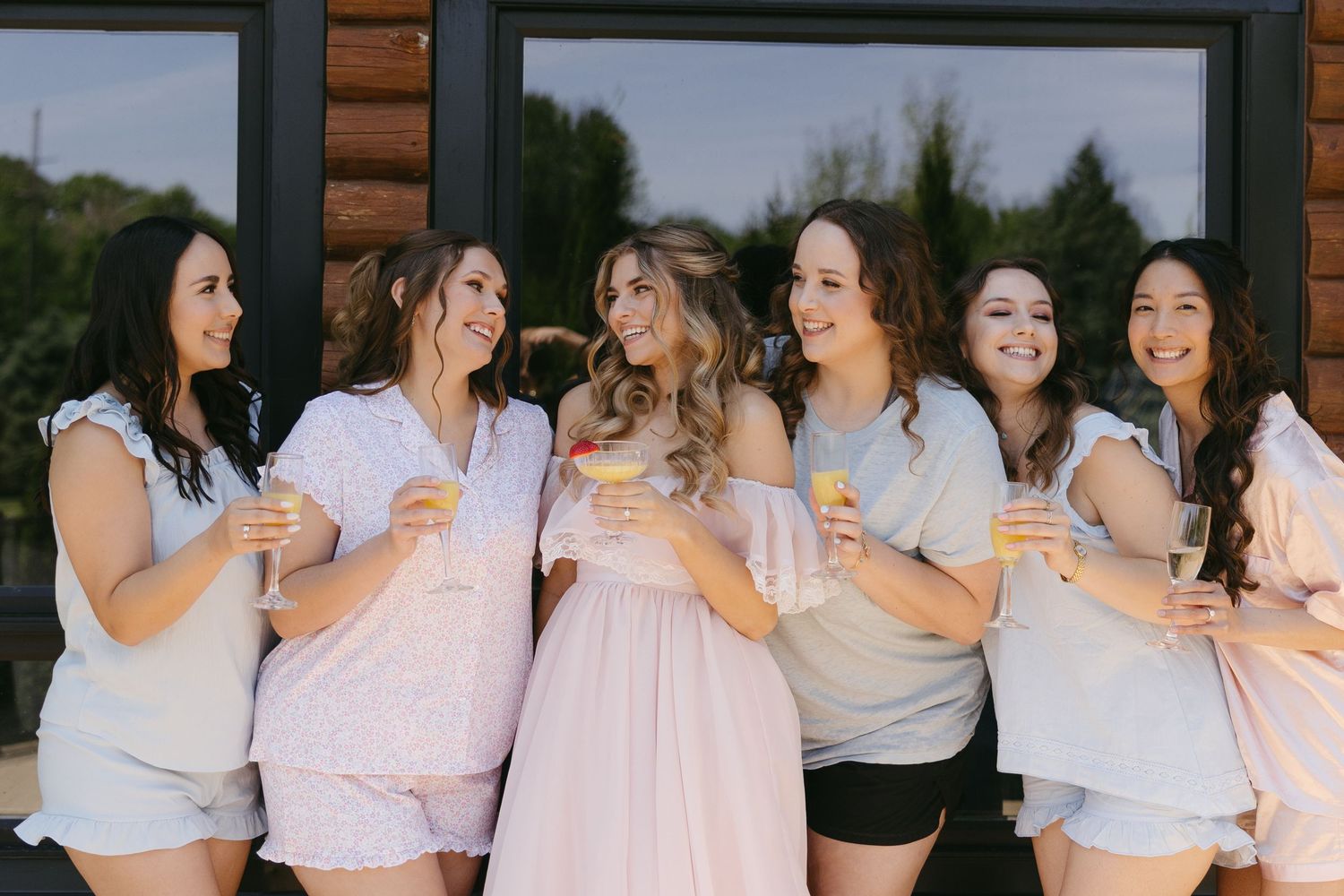 Six bridesmaids in pastel pajamas laughing and holding champagne glasses together during a fun bridal party getting ready celebration.