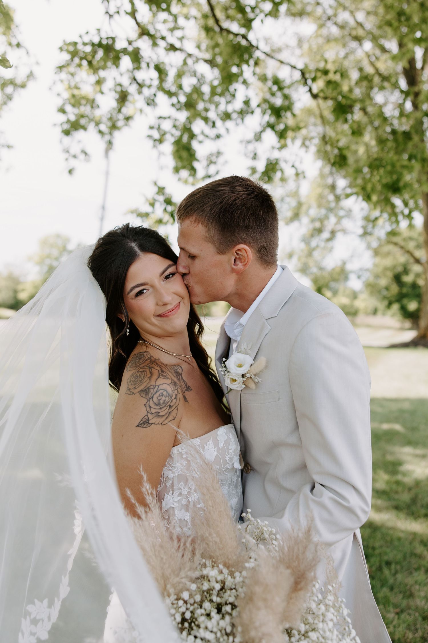 Groom in gray suit kisses tattooed bride on cheek as her veil flows in the breeze outdoors.