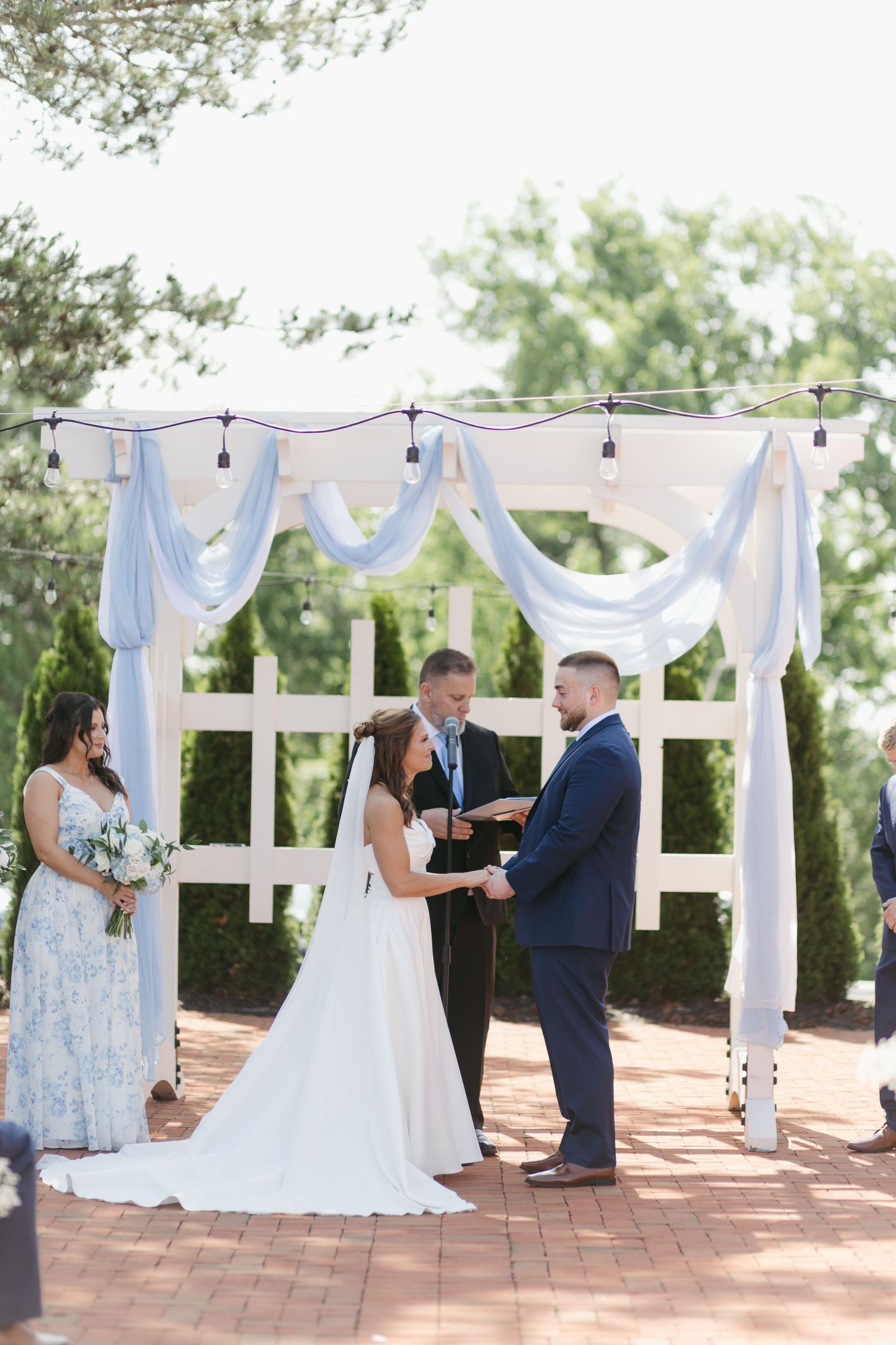Bride and groom exchange vows under a white arch draped in blue fabric at an outdoor ceremony.