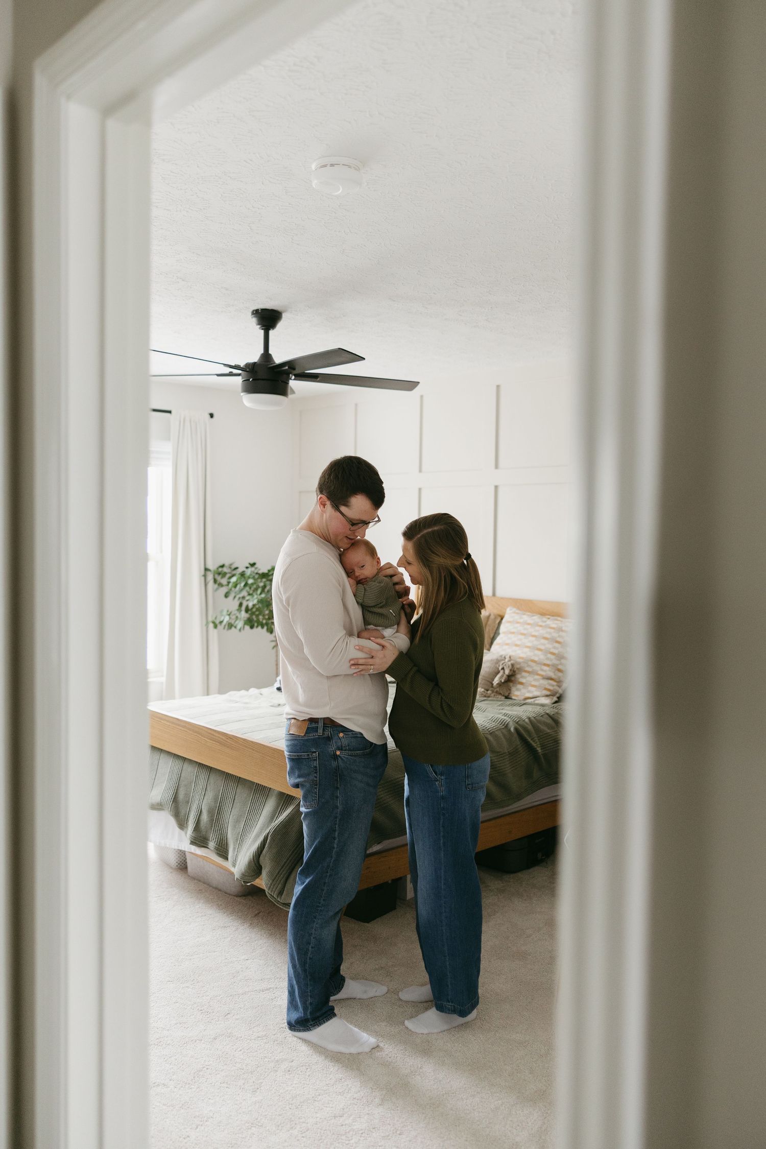 New parents stand in cozy bedroom doorway cradling their newborn baby together in warm home setting.