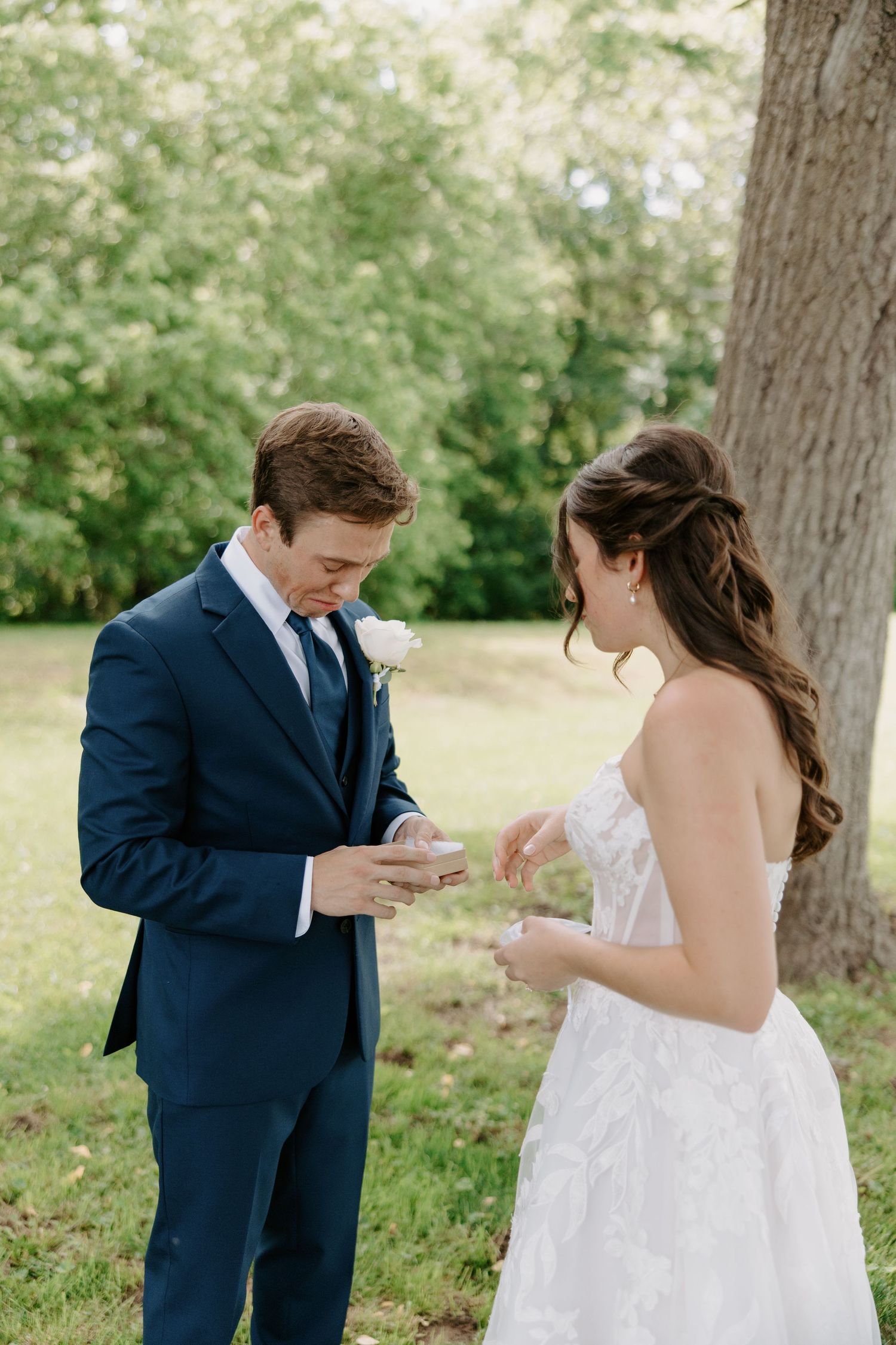 A groom in a navy suit and bride in a white gown exchange vows outdoors near a tree on their wedding day.
