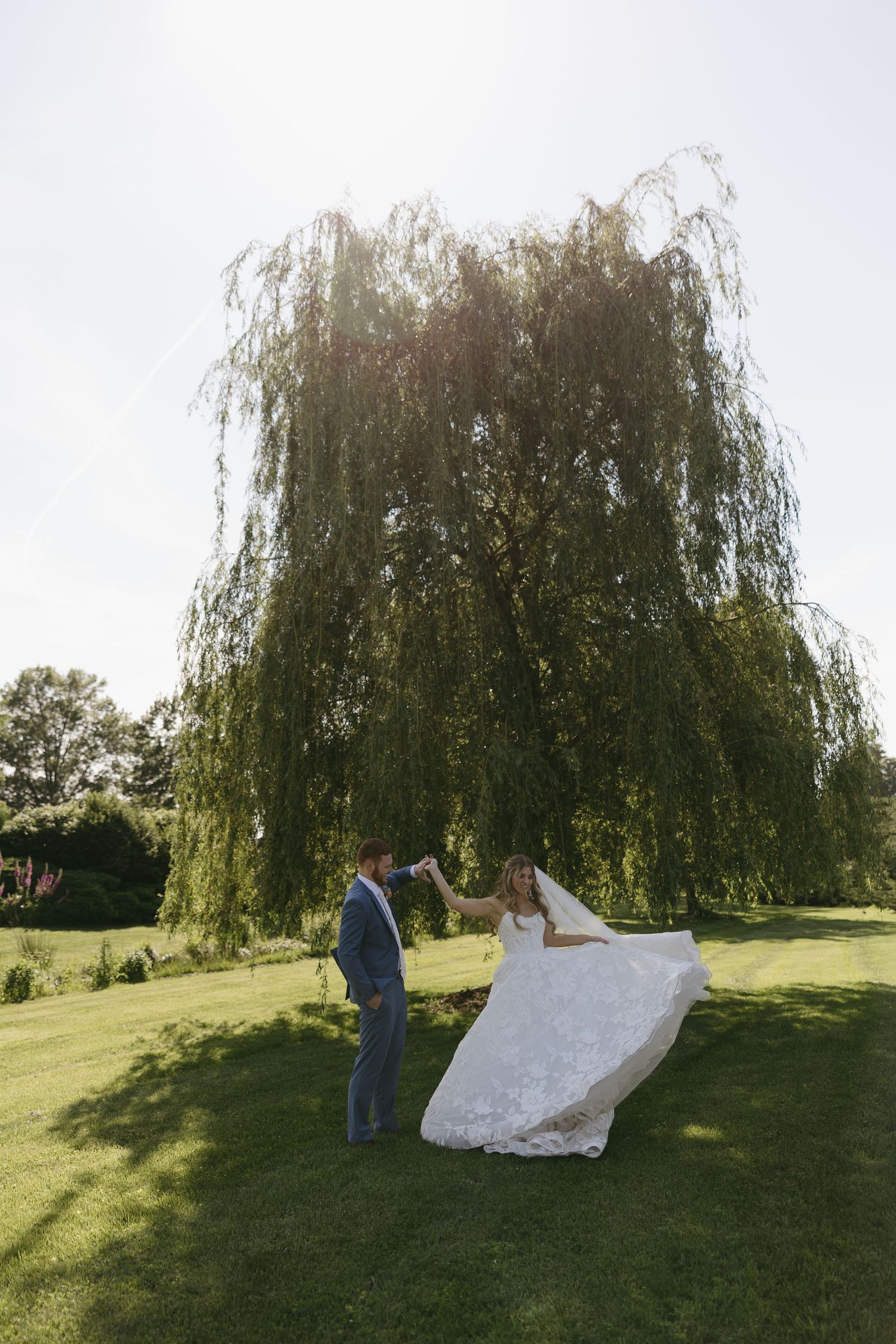 Bride and groom dancing on green lawn beneath a weeping willow tree on a sunny wedding day.