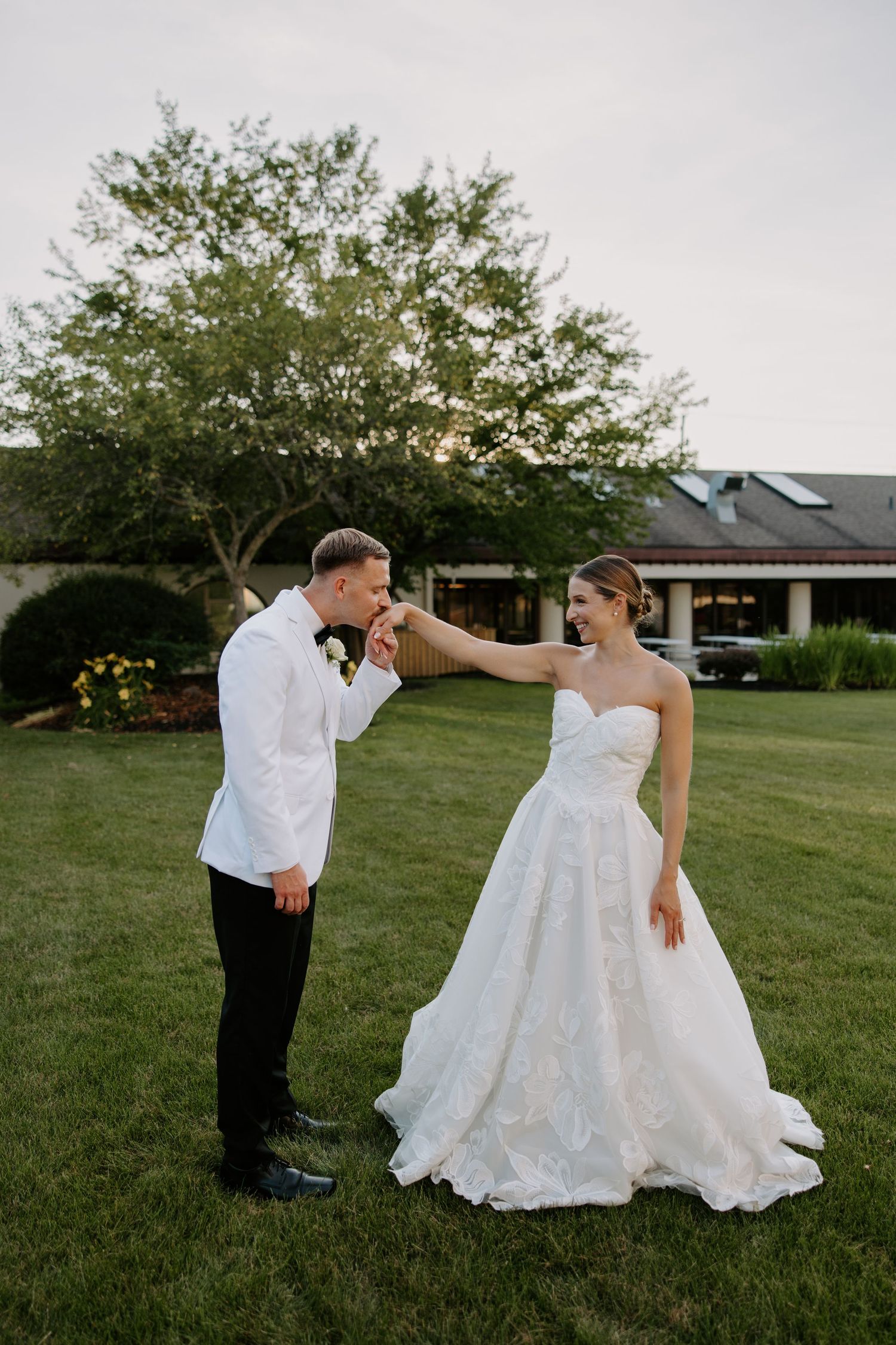 Groom in white jacket kissing bride's hand as she smiles in a strapless floral wedding gown on a lush green lawn.