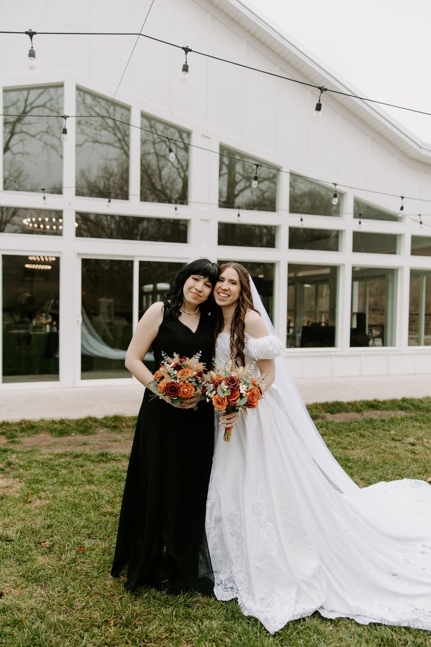 Bride in white ball gown and woman in black dress hold bouquets while posing outside bright white wedding venue.