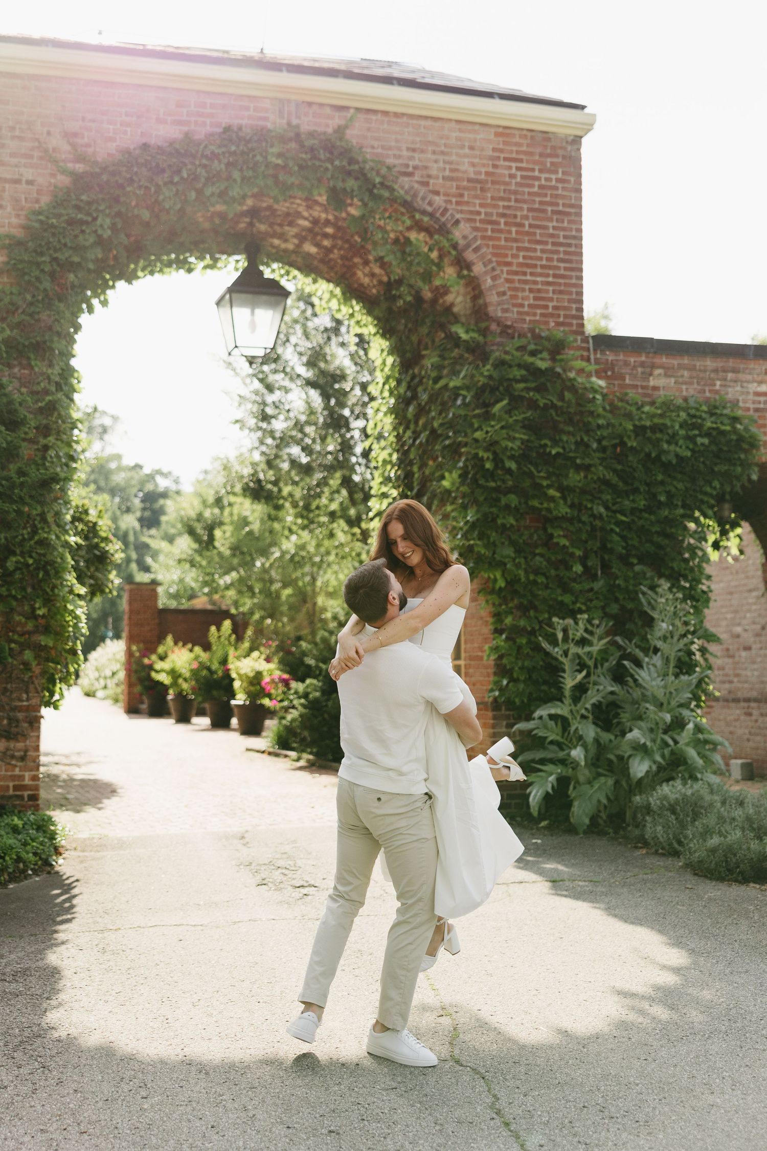 Groom lifts bride under an ivy-covered brick arch in a sunlit garden courtyard.