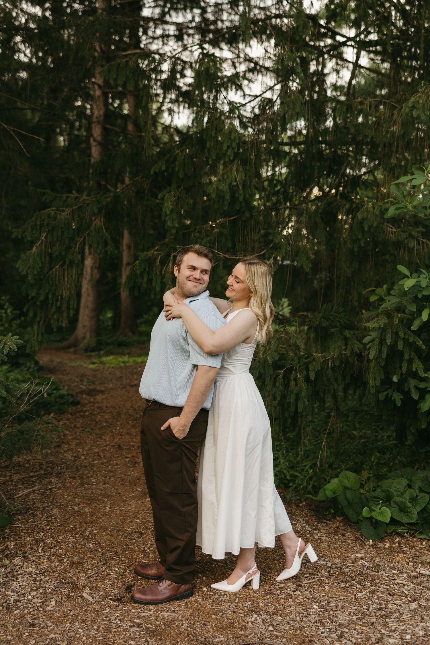 Couple embraces on a wooded gravel path surrounded by tall evergreen trees.