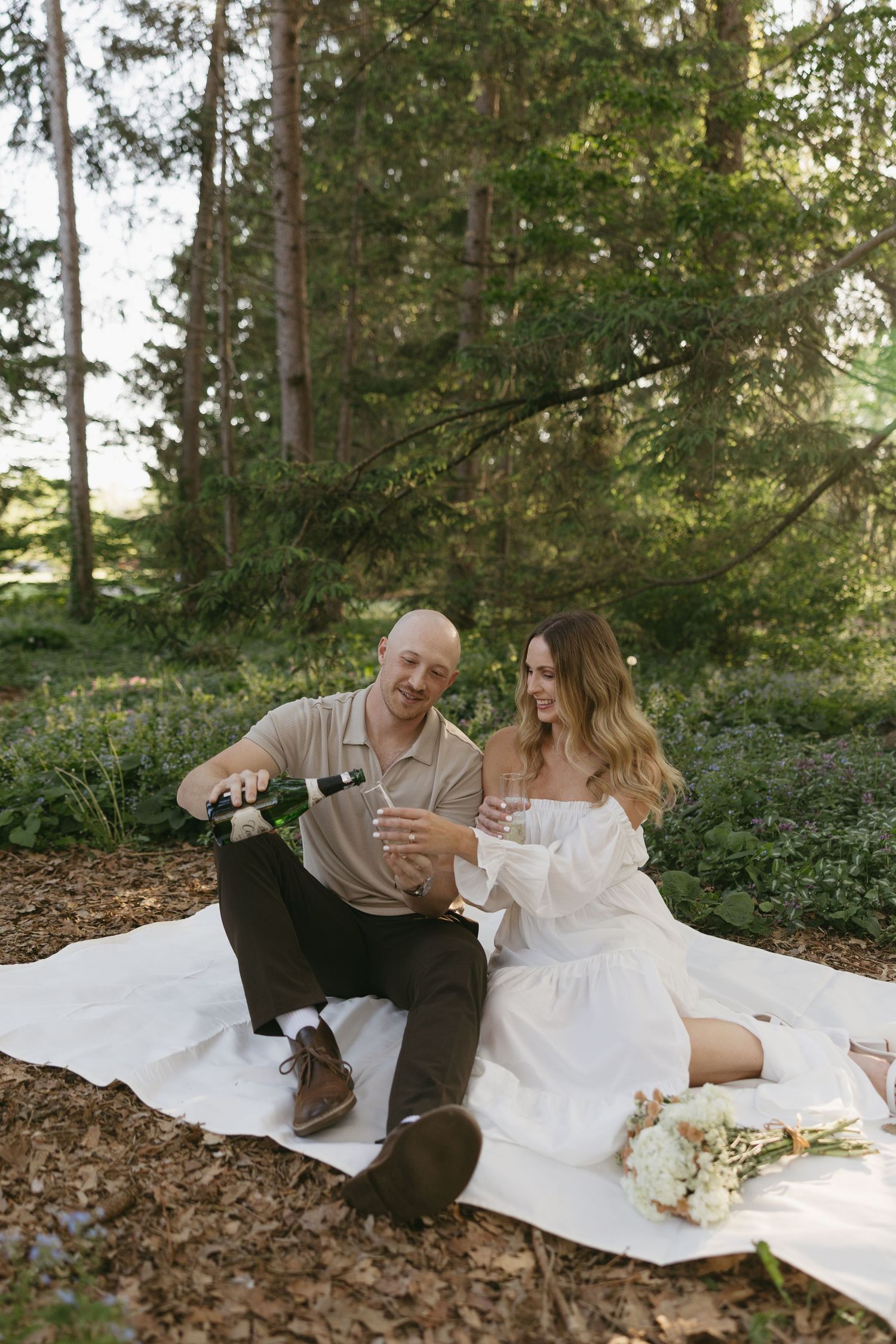 Couple sitting on a blanket in the woods, pouring champagne and laughing with a bouquet nearby.