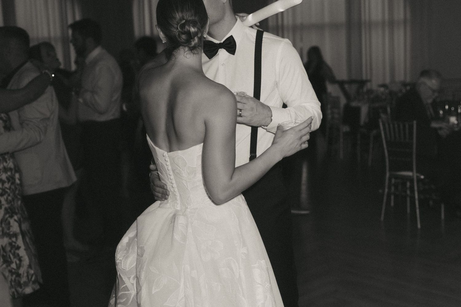 Black and white photo of bride and groom sharing first dance at wedding reception with guests in background.