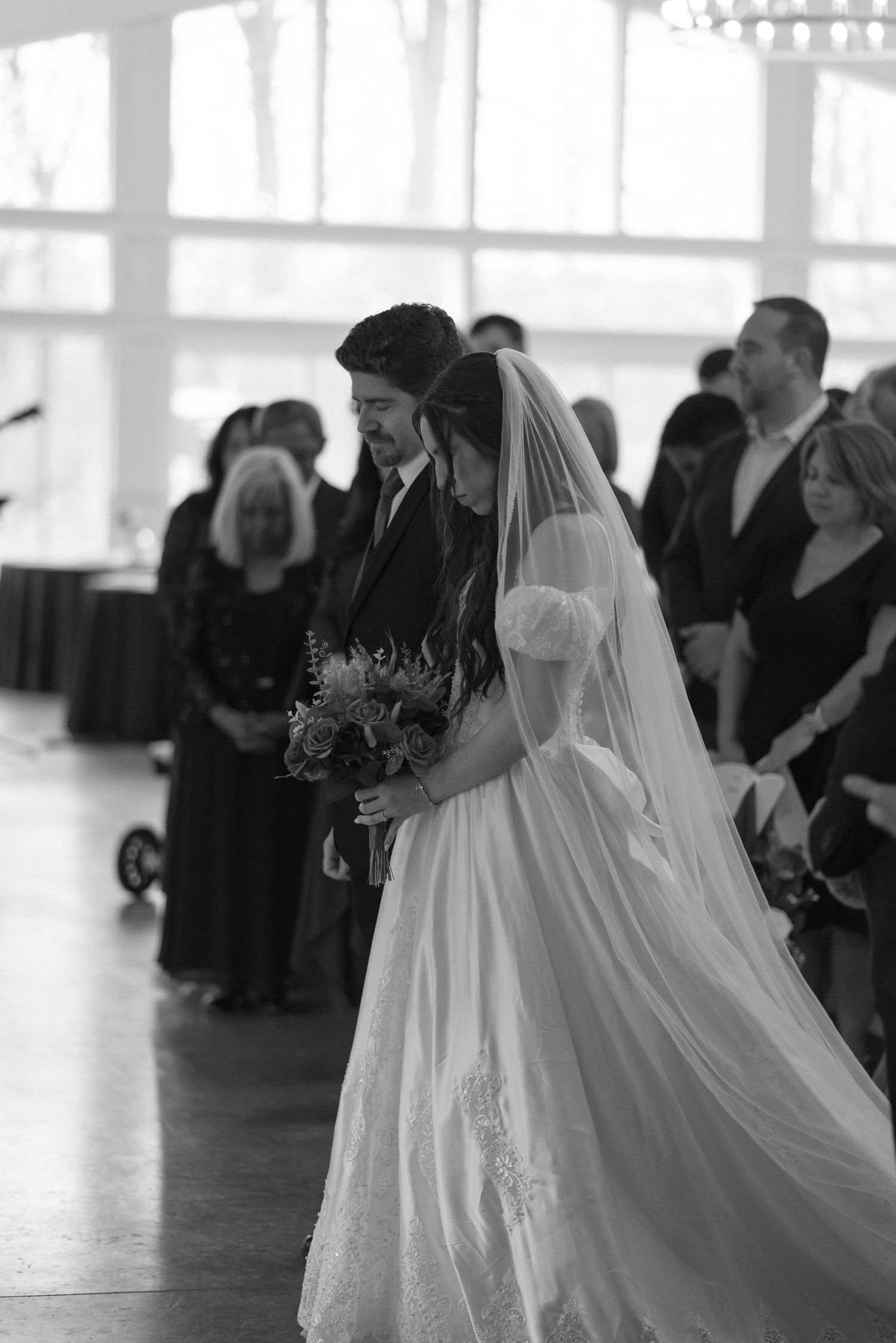 Black and white photo of bride and groom walking down aisle during indoor wedding ceremony with seated guests.