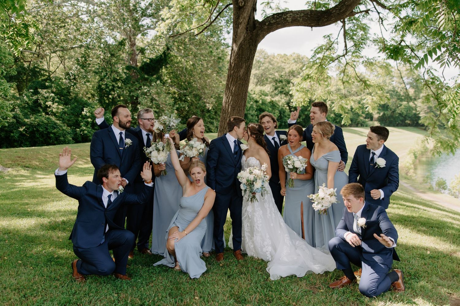 Wedding party celebrating under a tree outdoors, bridesmaids in dusty blue gowns with groomsmen in navy suits.