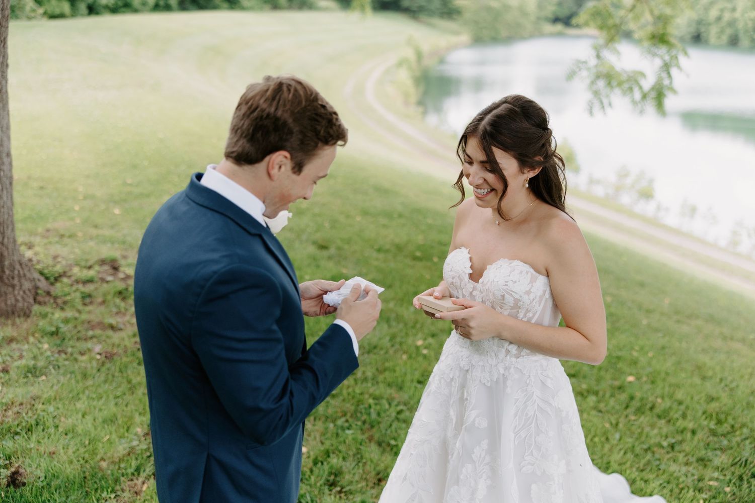 Bride and groom exchanging vows outdoors near a lake, dressed in a white lace gown and navy suit.