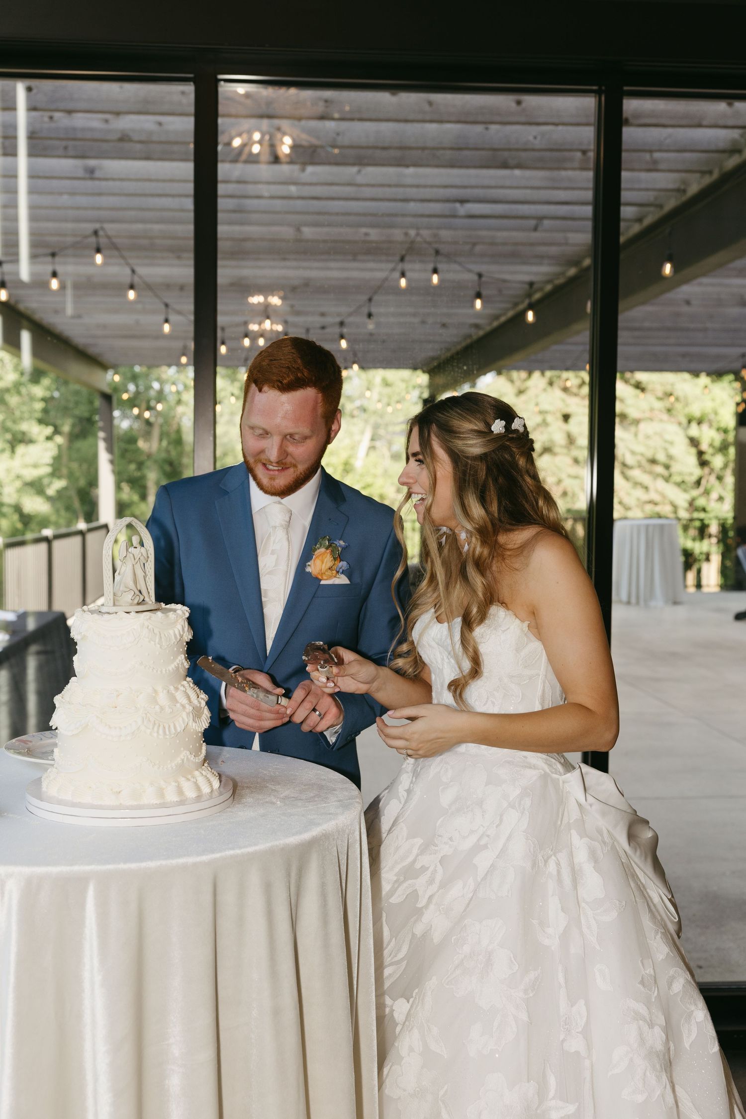 Bride and groom laughing while cutting white wedding cake at elegant indoor reception venue with string lights.