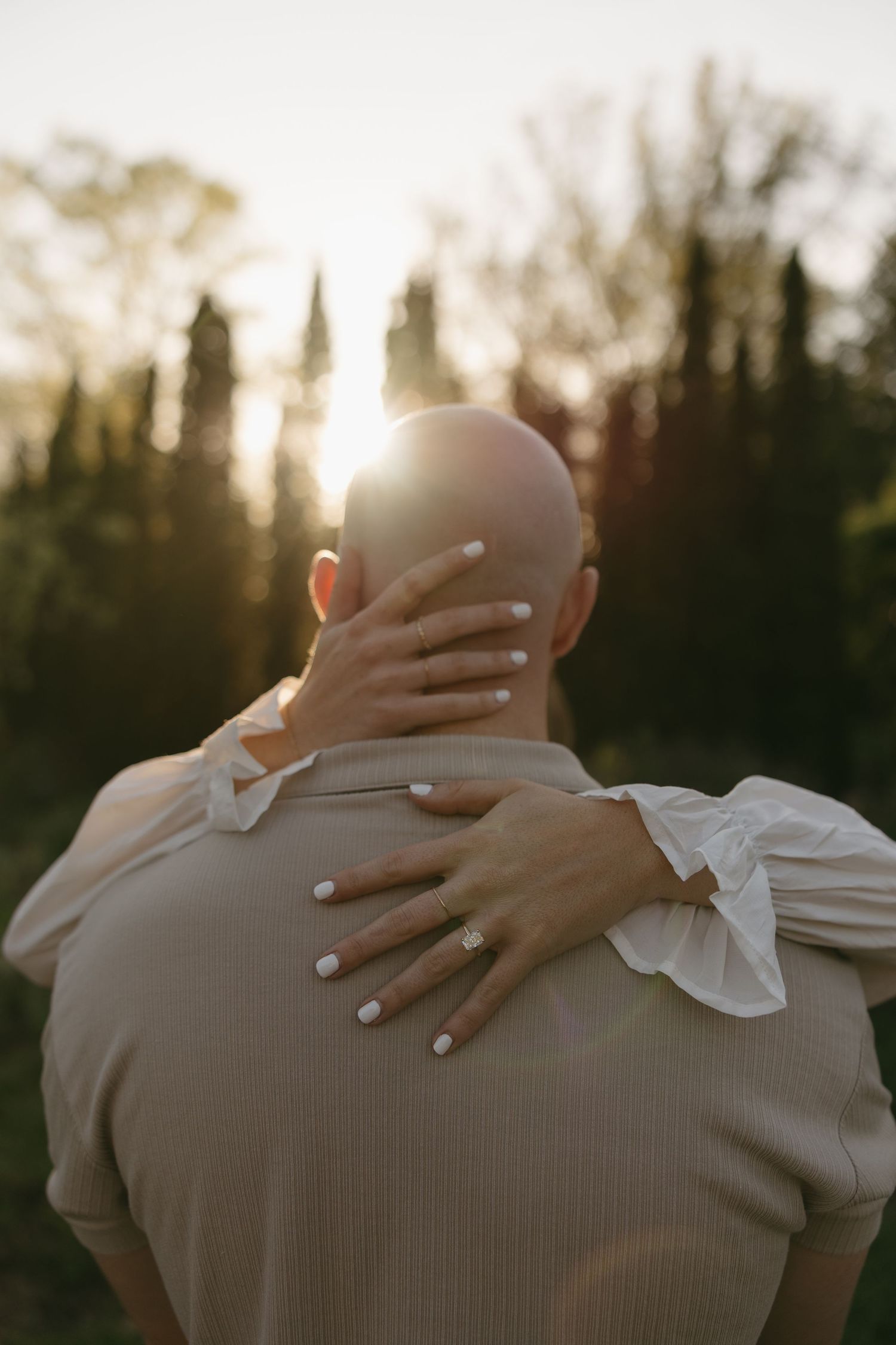 Back view of woman placing hands on bald mans shoulders with an engagement ring visible at sunset.