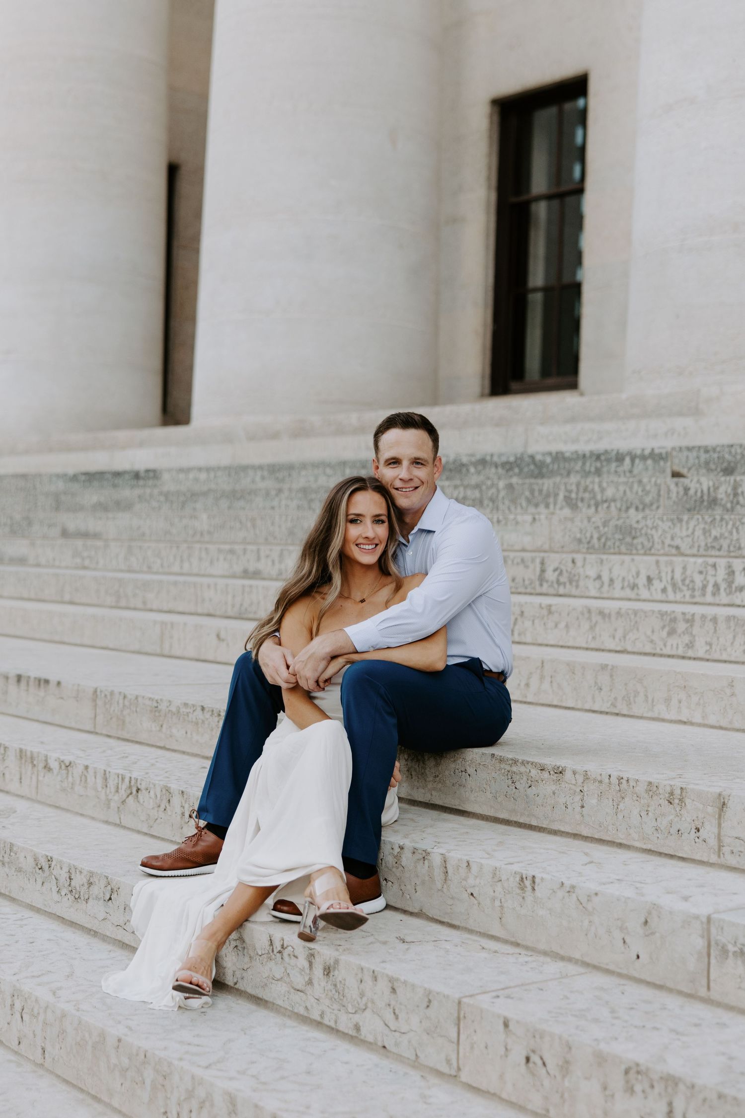 Engaged couple sitting together on marble steps of a classical building, smiling at the camera.