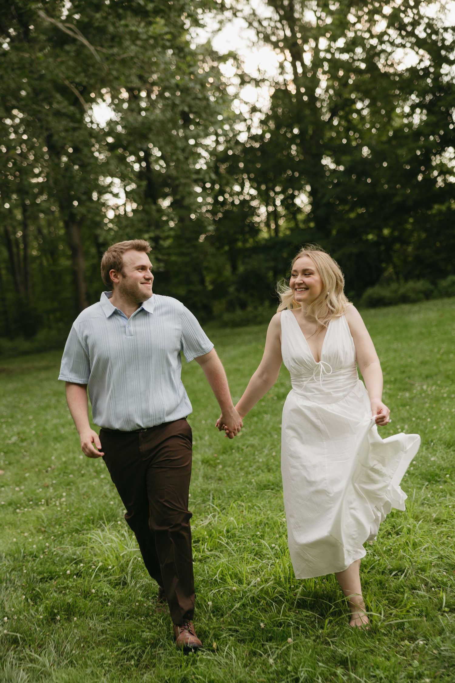 Happy couple holding hands and walking through a green meadow surrounded by trees at golden hour.