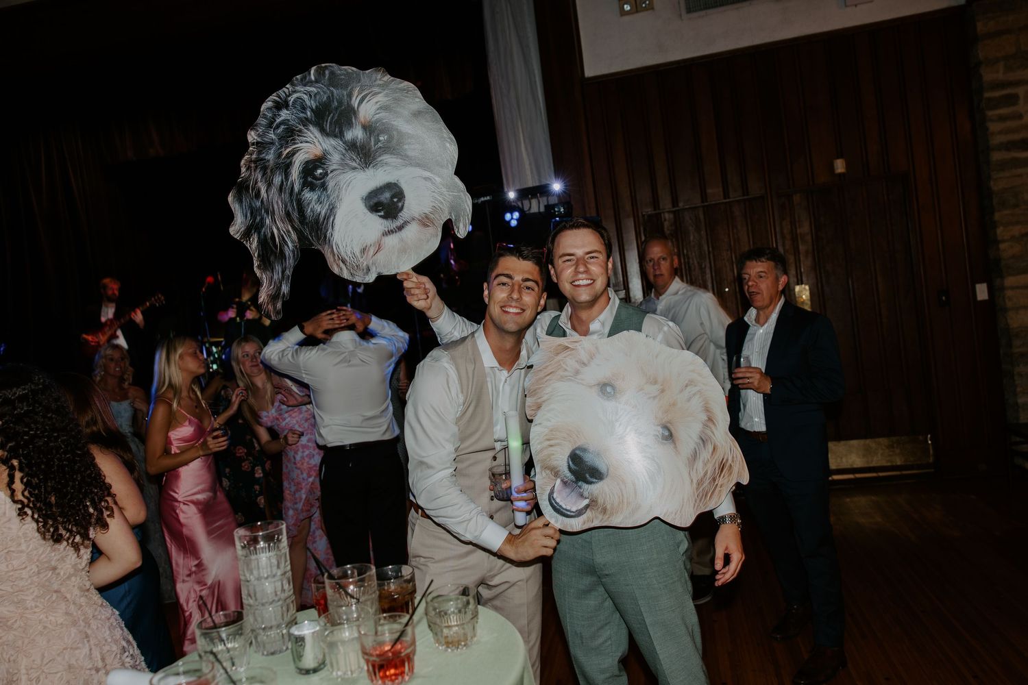 Two men pose with oversized dog face cutouts at a lively wedding reception party.