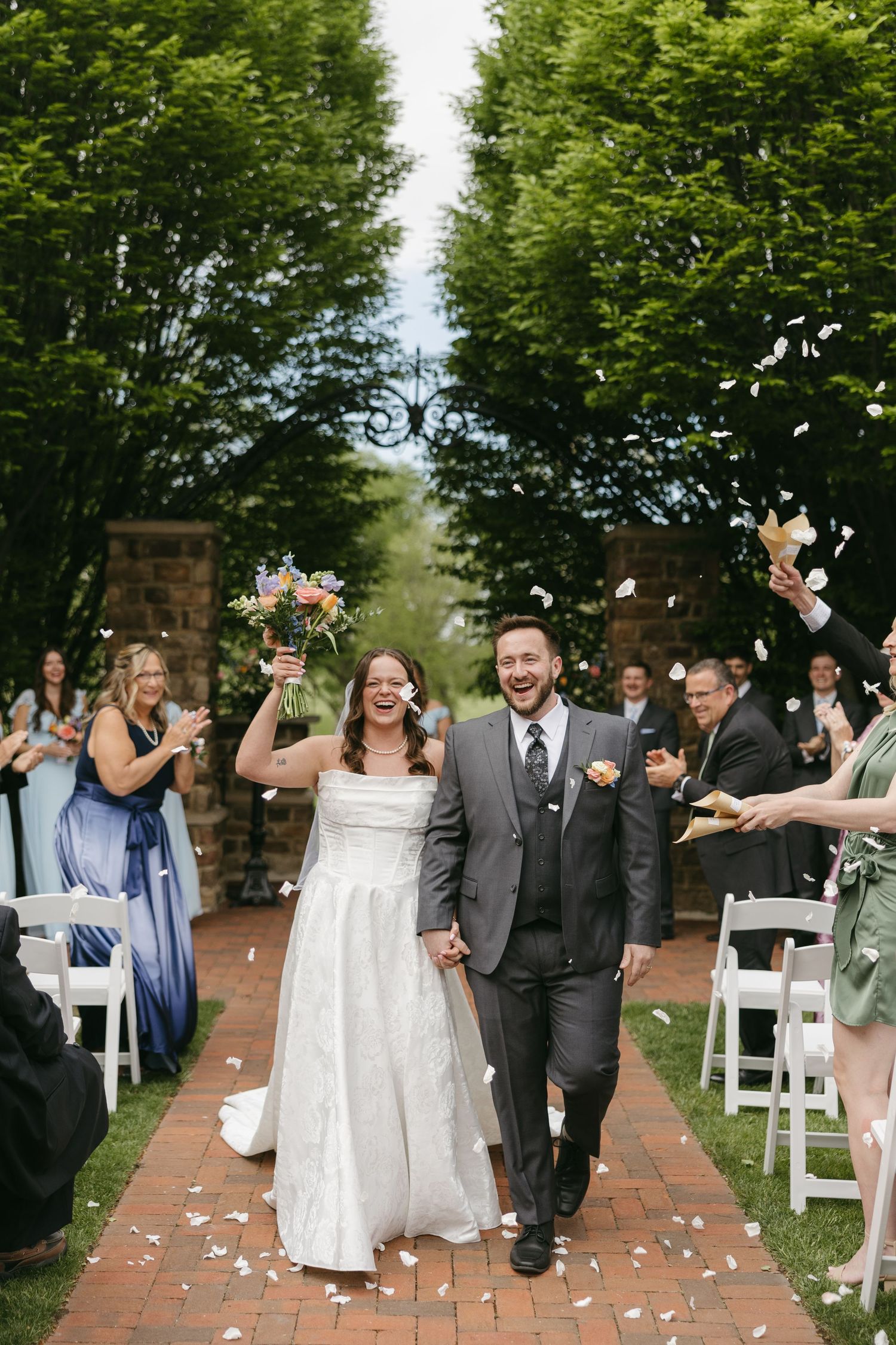 Newlywed couple walks joyfully down petal-lined aisle as guests toss flower petals in outdoor ceremony.