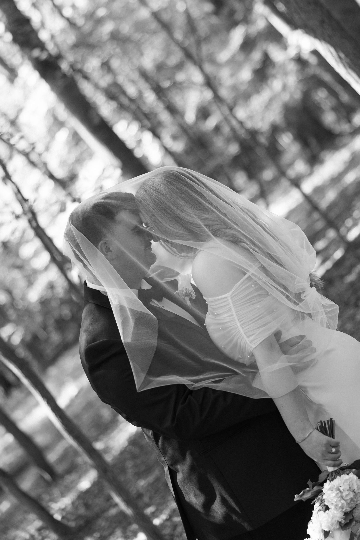 Black and white romantic photo of bride and groom kissing under a flowing veil in a forest.