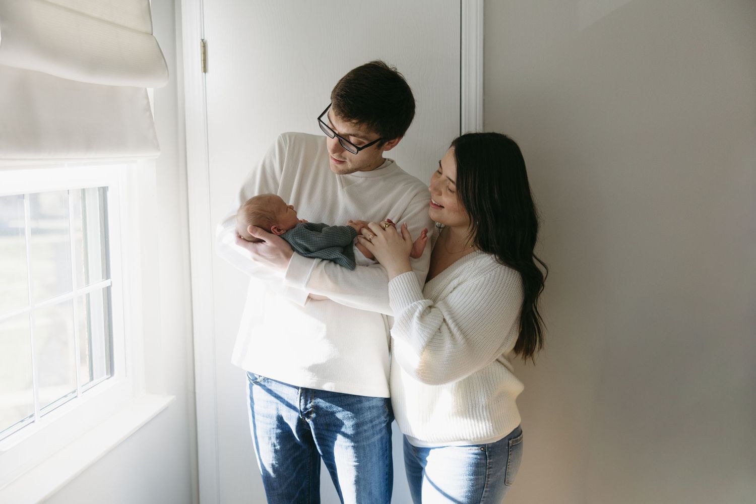 Happy young couple standing by a window, lovingly holding their newborn baby together.
