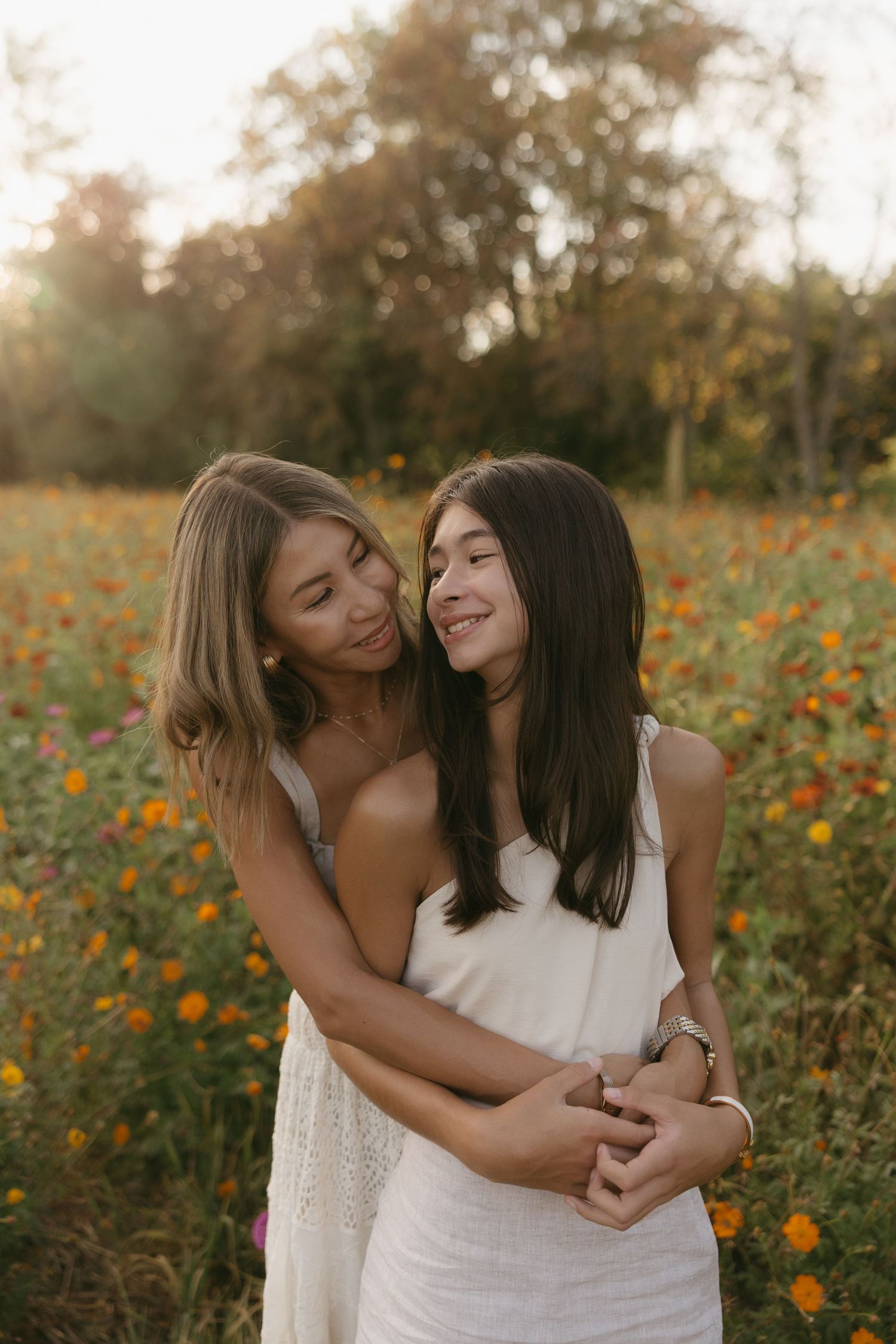 Mother and teenage daughter embracing and smiling in a colorful flower field at sunset.