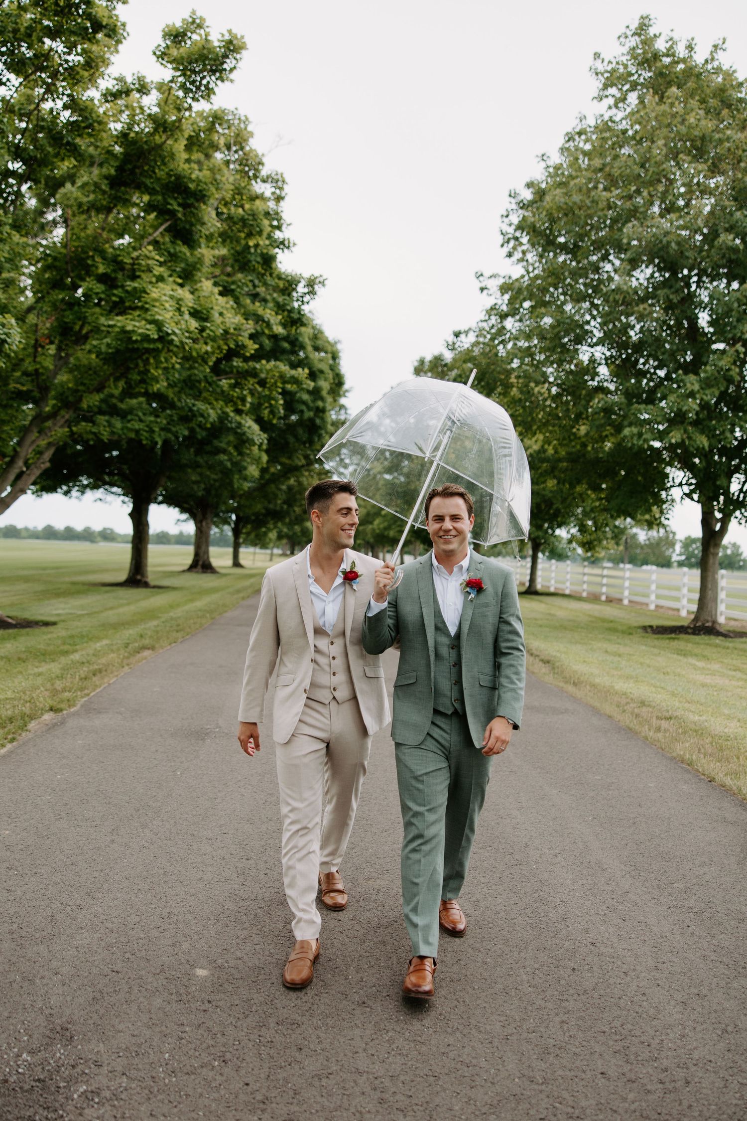 Two grooms in suits walk under a clear bubble umbrella on a tree-lined path at their outdoor wedding.