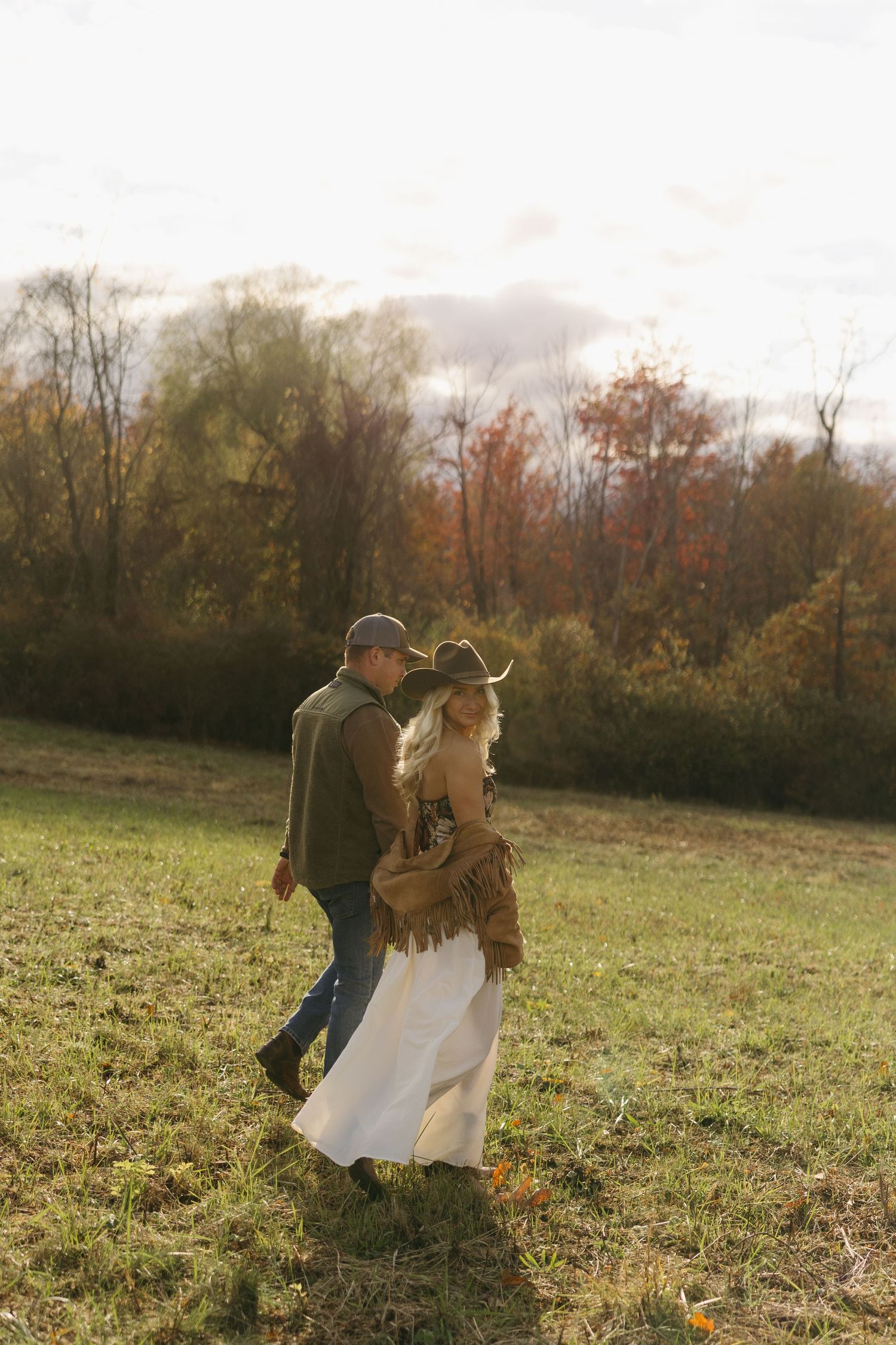 Couple walking through an autumn field, woman in white dress and cowboy hat, man in green jacket, colorful fall foliage backdrop.