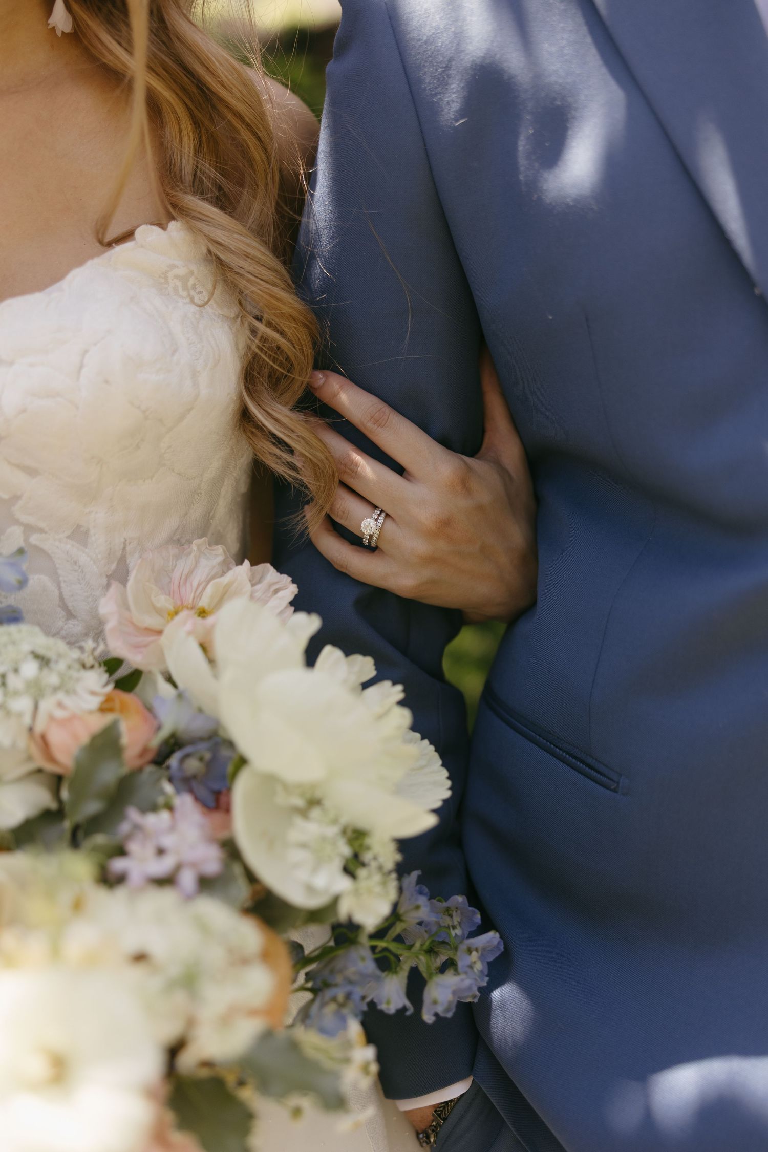 Close-up of bride holding colorful bouquet while resting hand on grooms blue suit showing wedding ring.