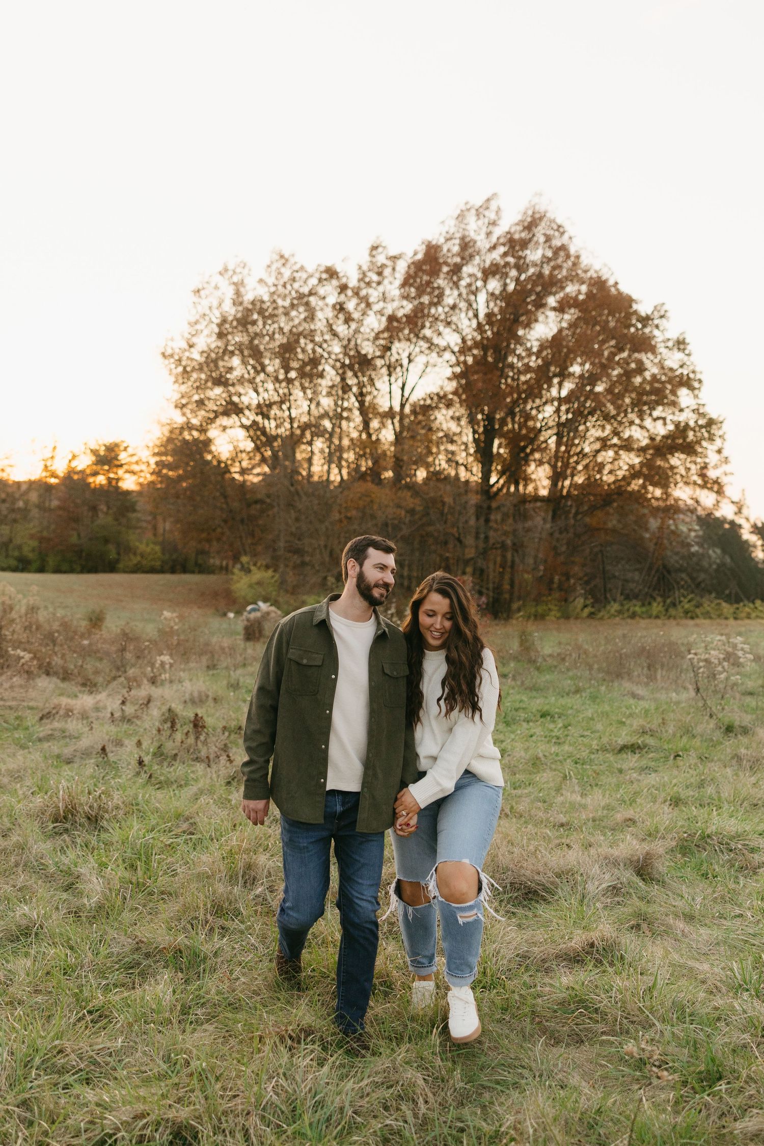 Couple walking hand in hand through an open meadow at sunset, man in green jacket and woman in ripped jeans.