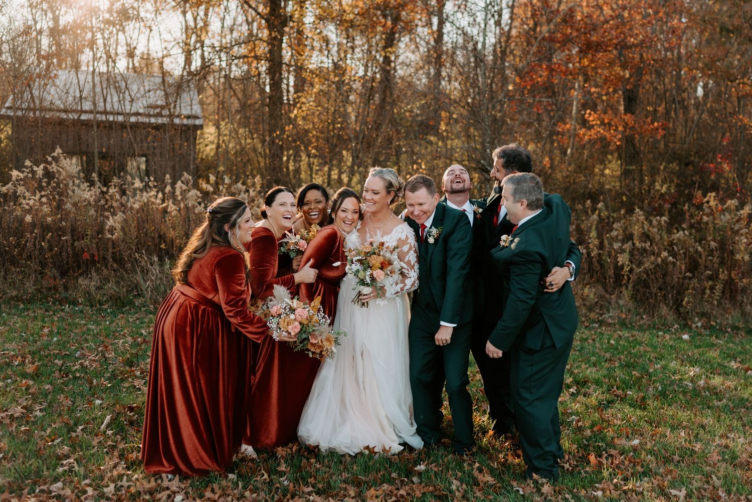 Bride with autumn bouquet stands with wedding party in rich red velvet and dark green suits among fall foliage.