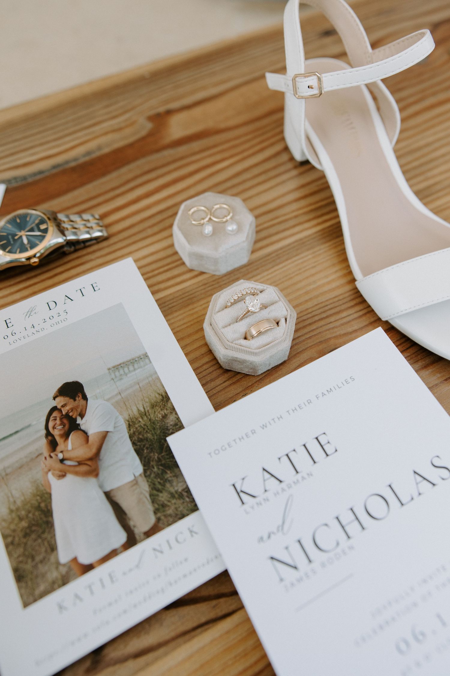 Elegant wedding flat lay featuring Katie and Nicholas invitation suite, gold rings in velvet box, white heels, and a watch on wood.