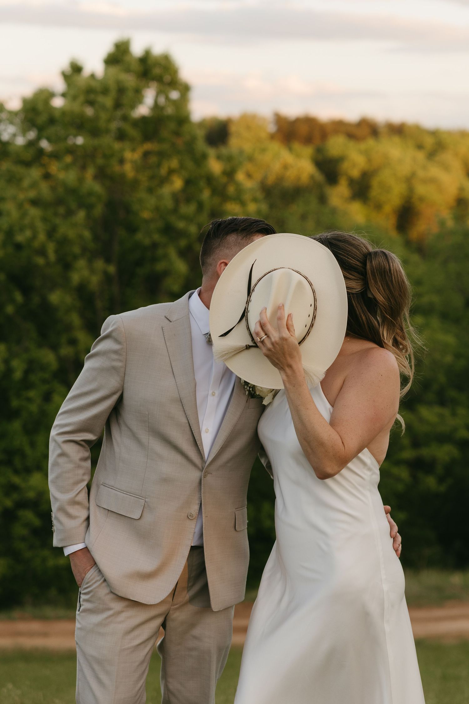 Bride and groom sharing a kiss hidden behind a wide-brim straw hat at golden hour outdoors.