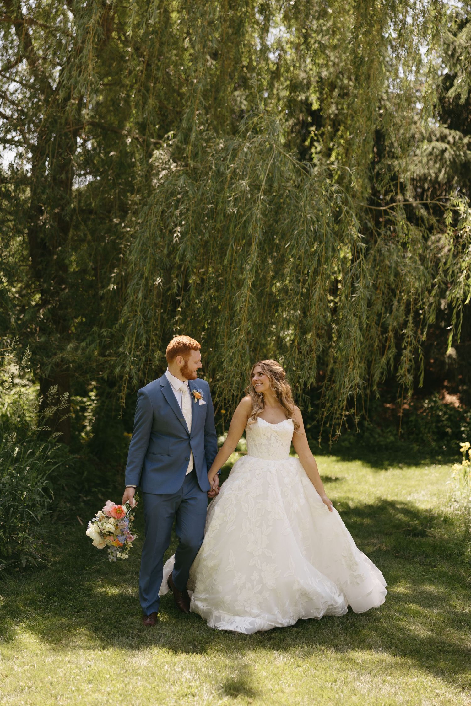Bride and groom walk hand-in-hand on green lawn beneath a weeping willow tree on their wedding day.