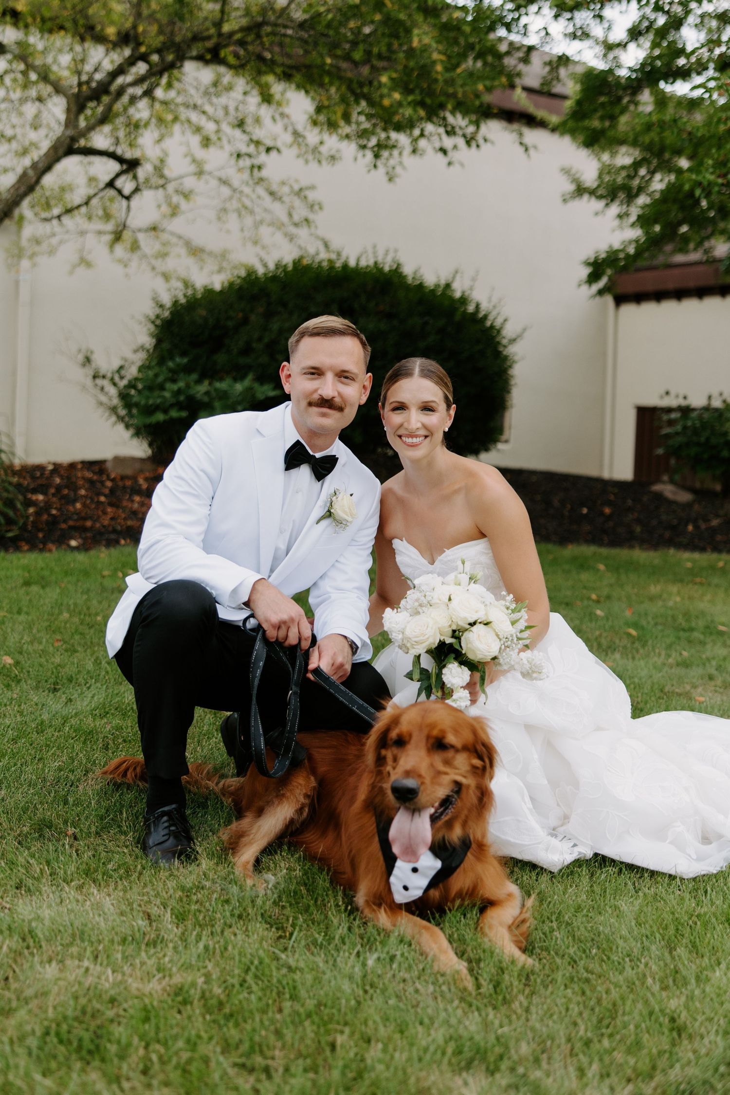 Bride and groom pose with their golden retriever dog wearing a tuxedo bow tie on the lawn.