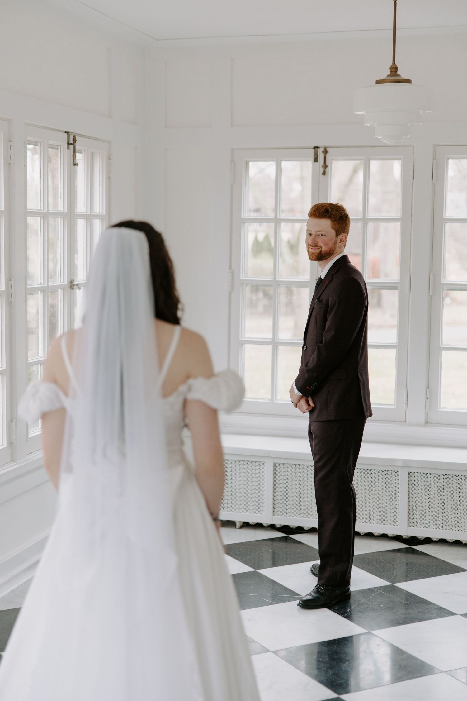 Groom in dark suit seeing bride in white off-shoulder gown and veil for first look on checkered floor.