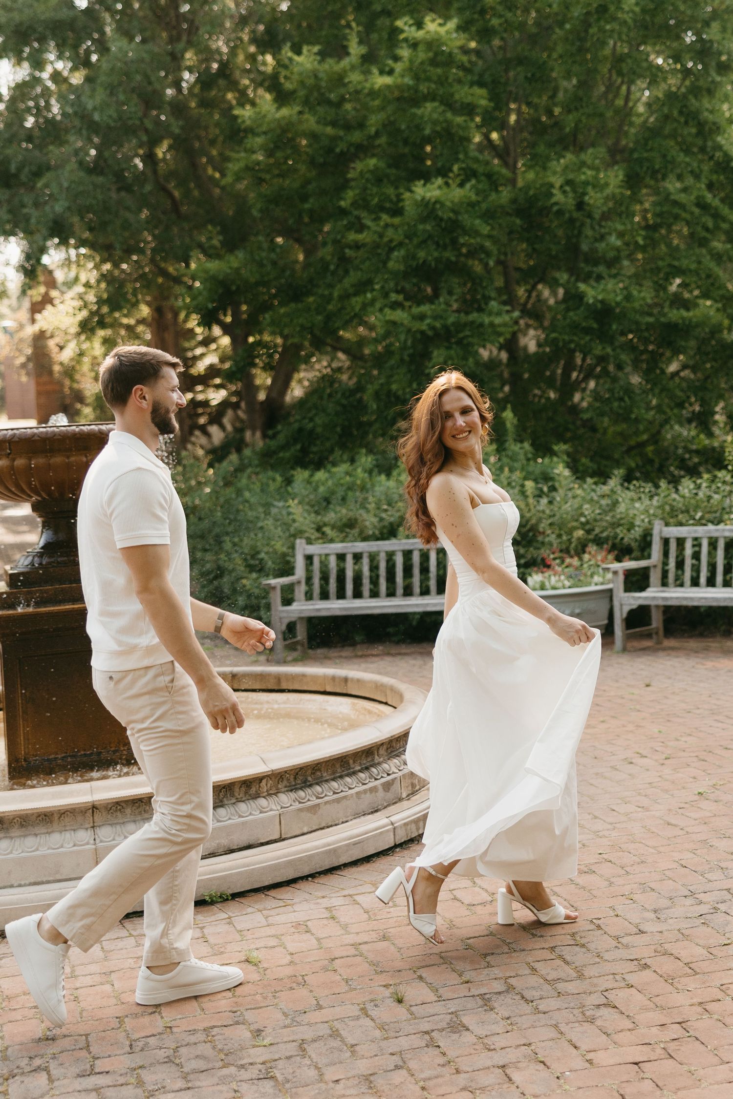 Bride in white dress and groom in beige outfit hold hands walking by a stone fountain in a lush garden.