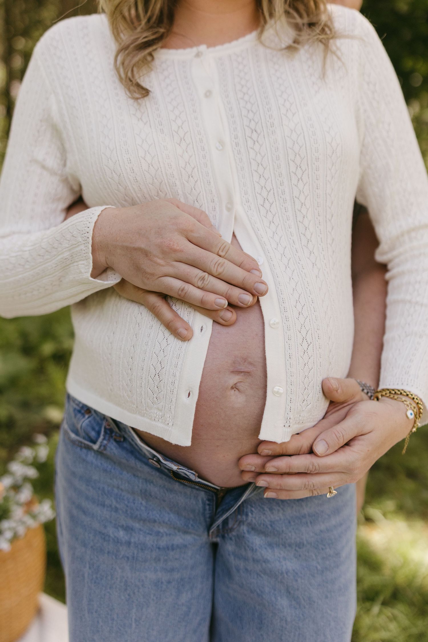 Close-up of a pregnant woman's belly being cradled by both partners' hands outdoors.