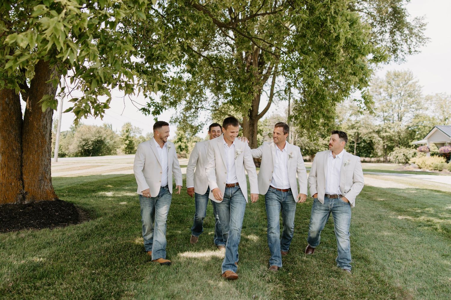 Groom and groomsmen in white blazers and jeans walk together under a shady tree outdoors.