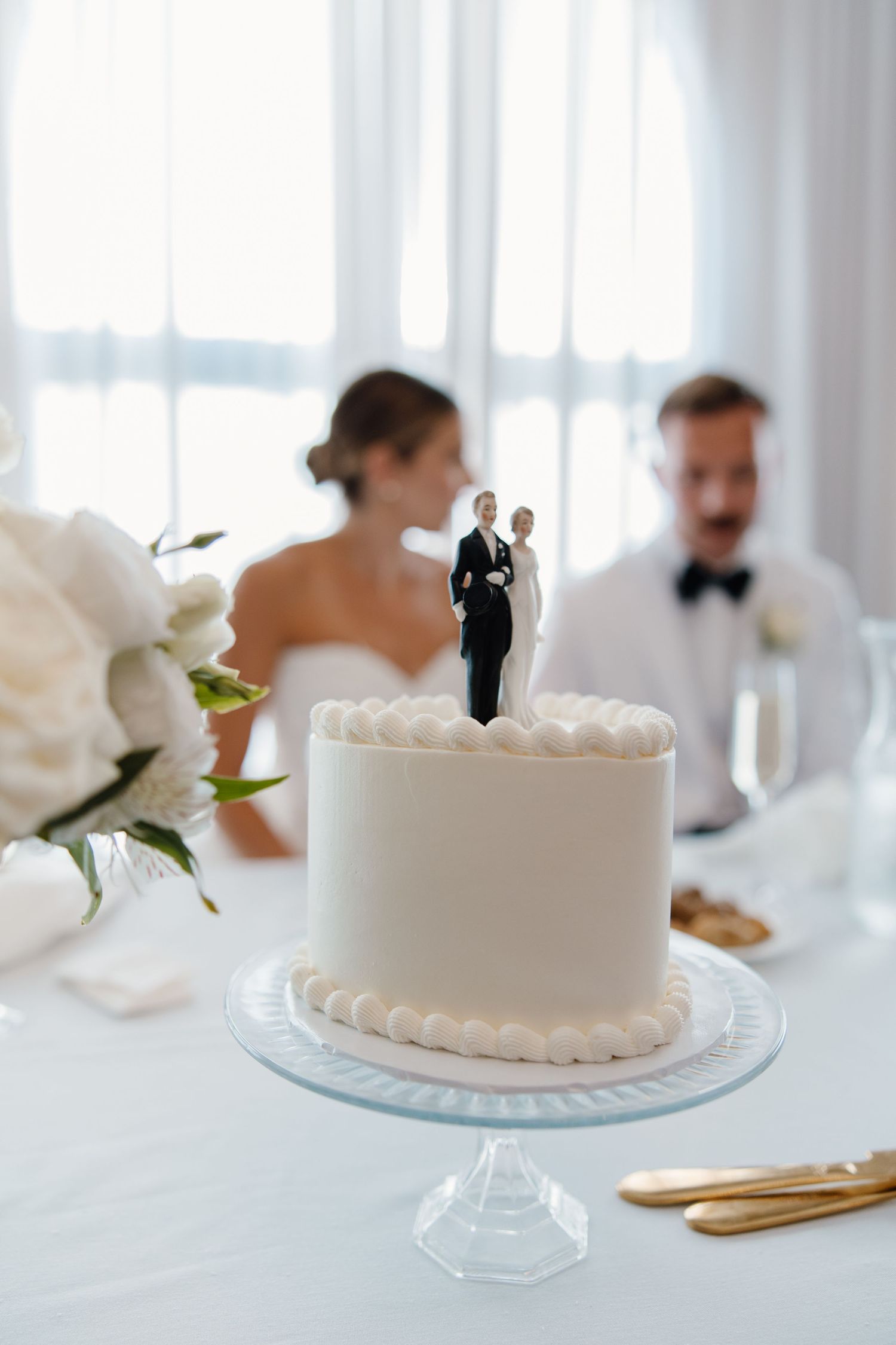 Simple white wedding cake with classic bride and groom topper displayed at elegant reception table.