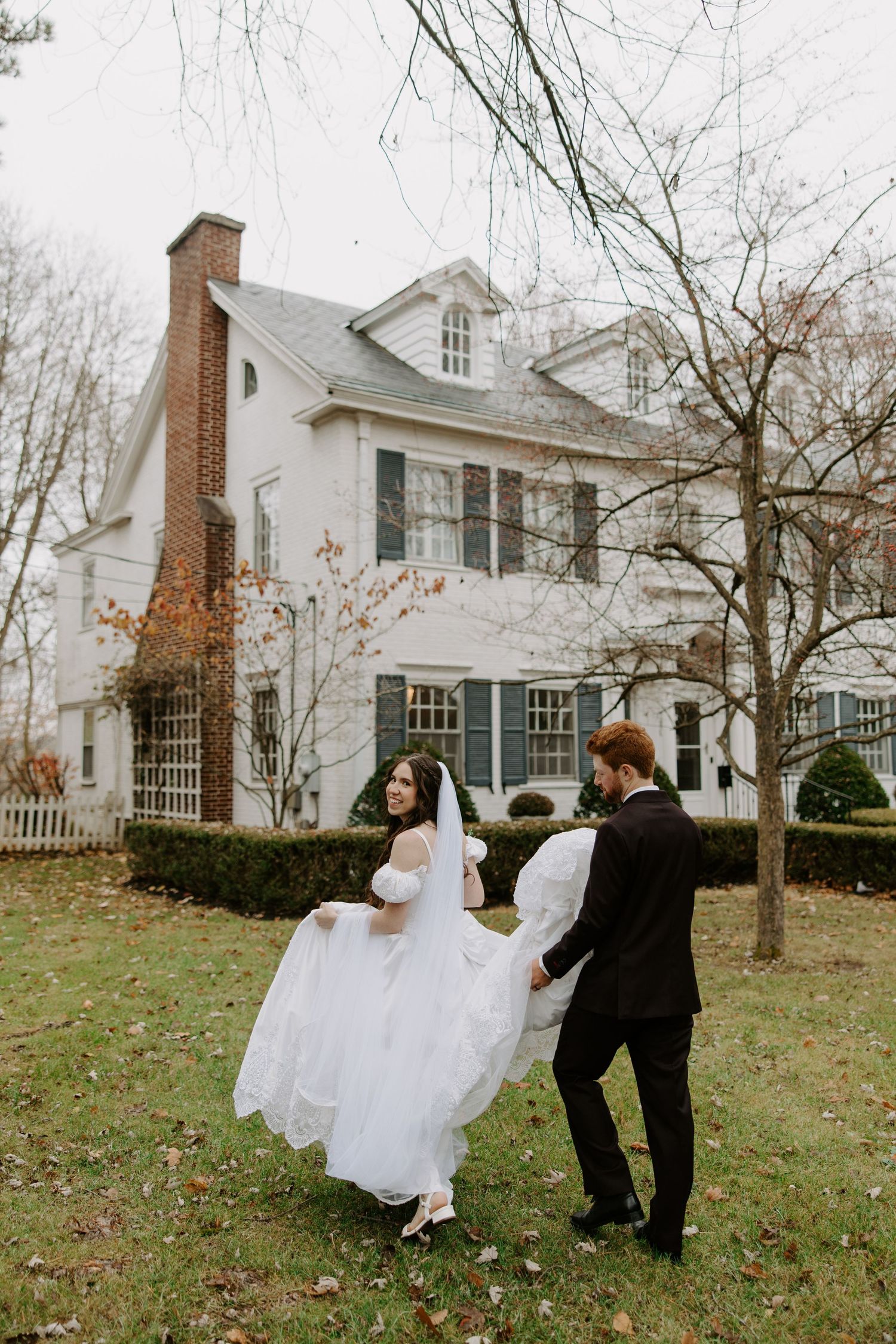 Bride in puff-sleeve gown with long veil twirling with groom in dark suit outside a white colonial estate.