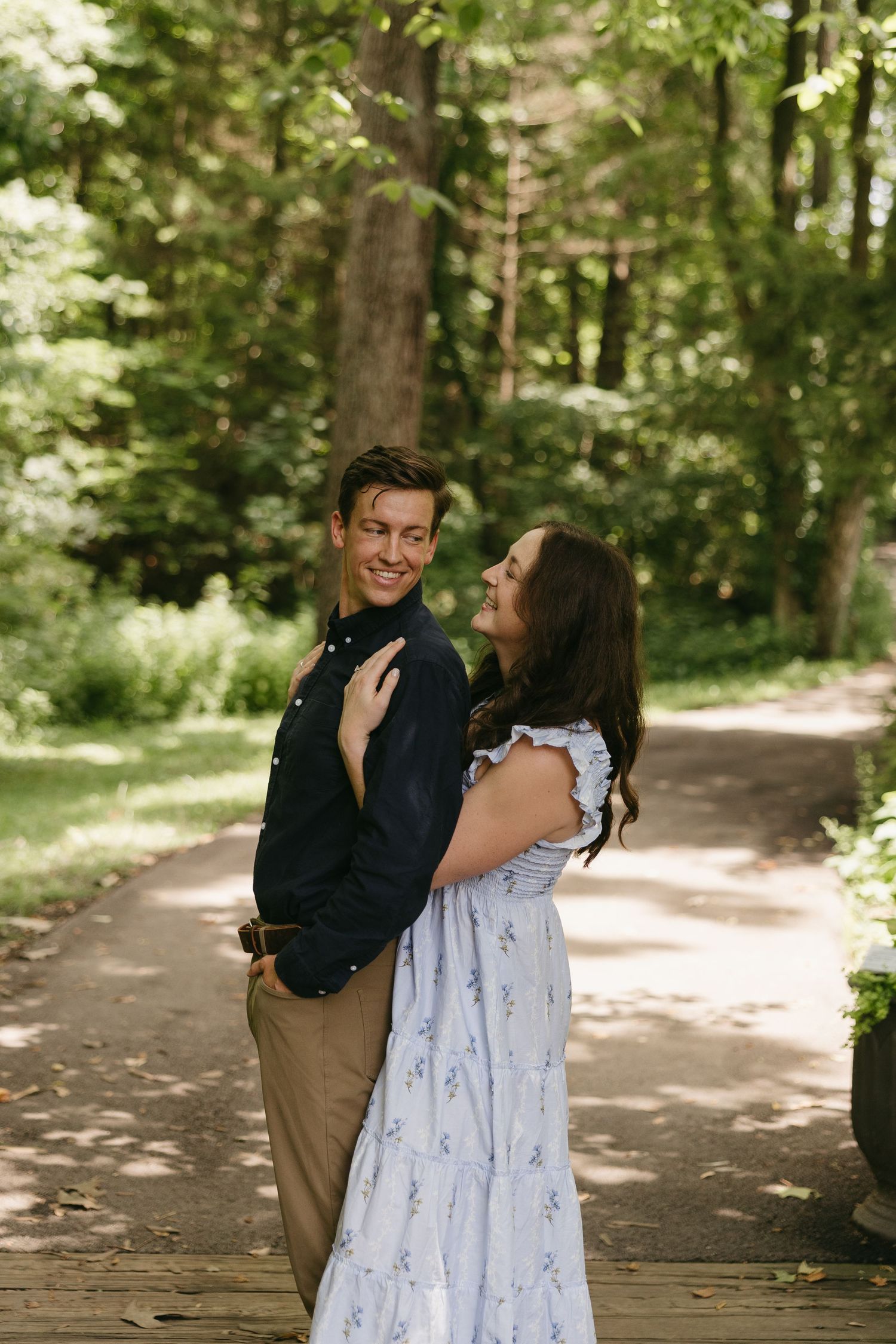 Couple embracing and smiling at each other on a shaded woodland path surrounded by green trees.