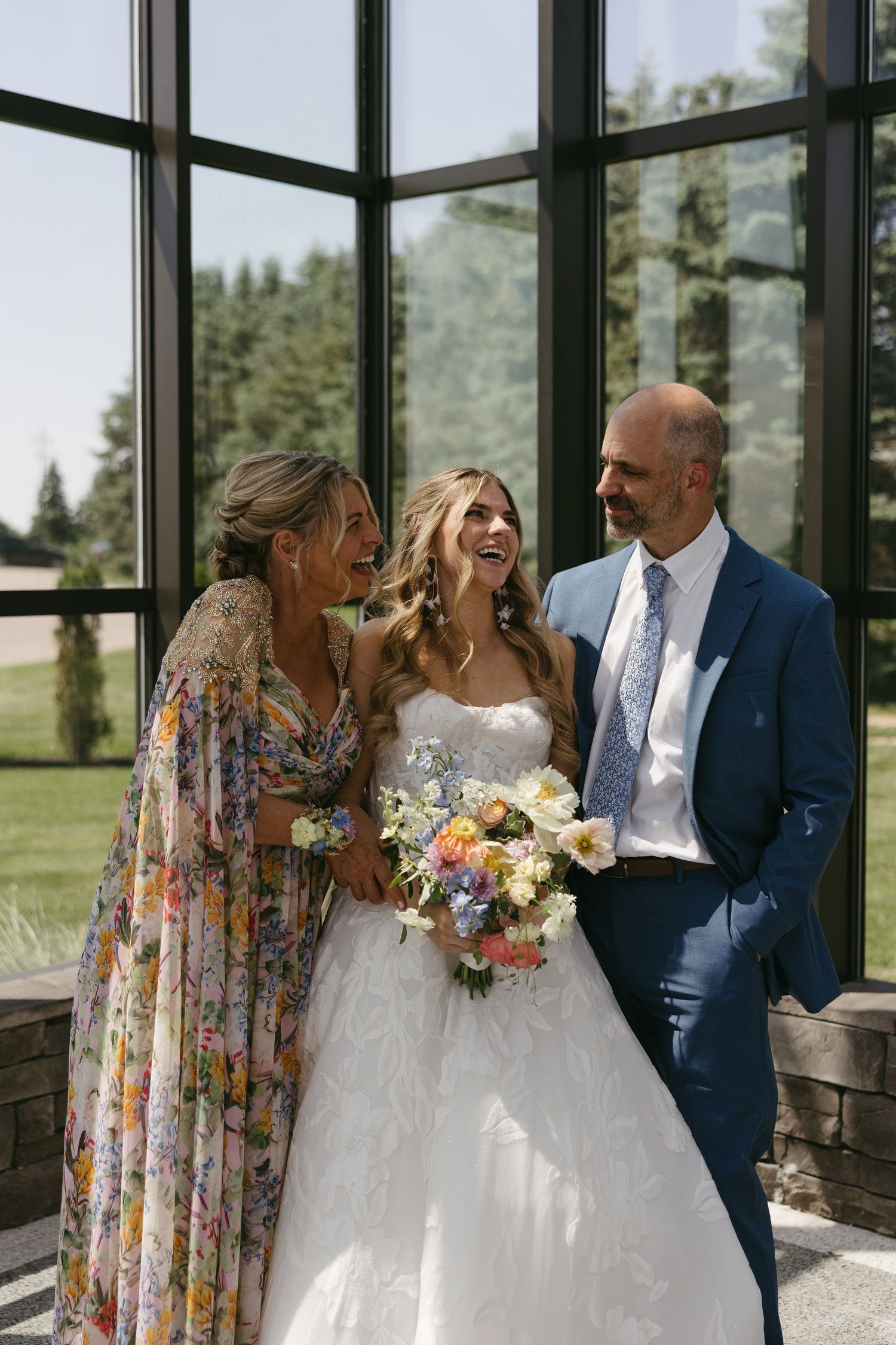 Bride in white gown laughing with parents in floral dress and blue suit near large windows at wedding.