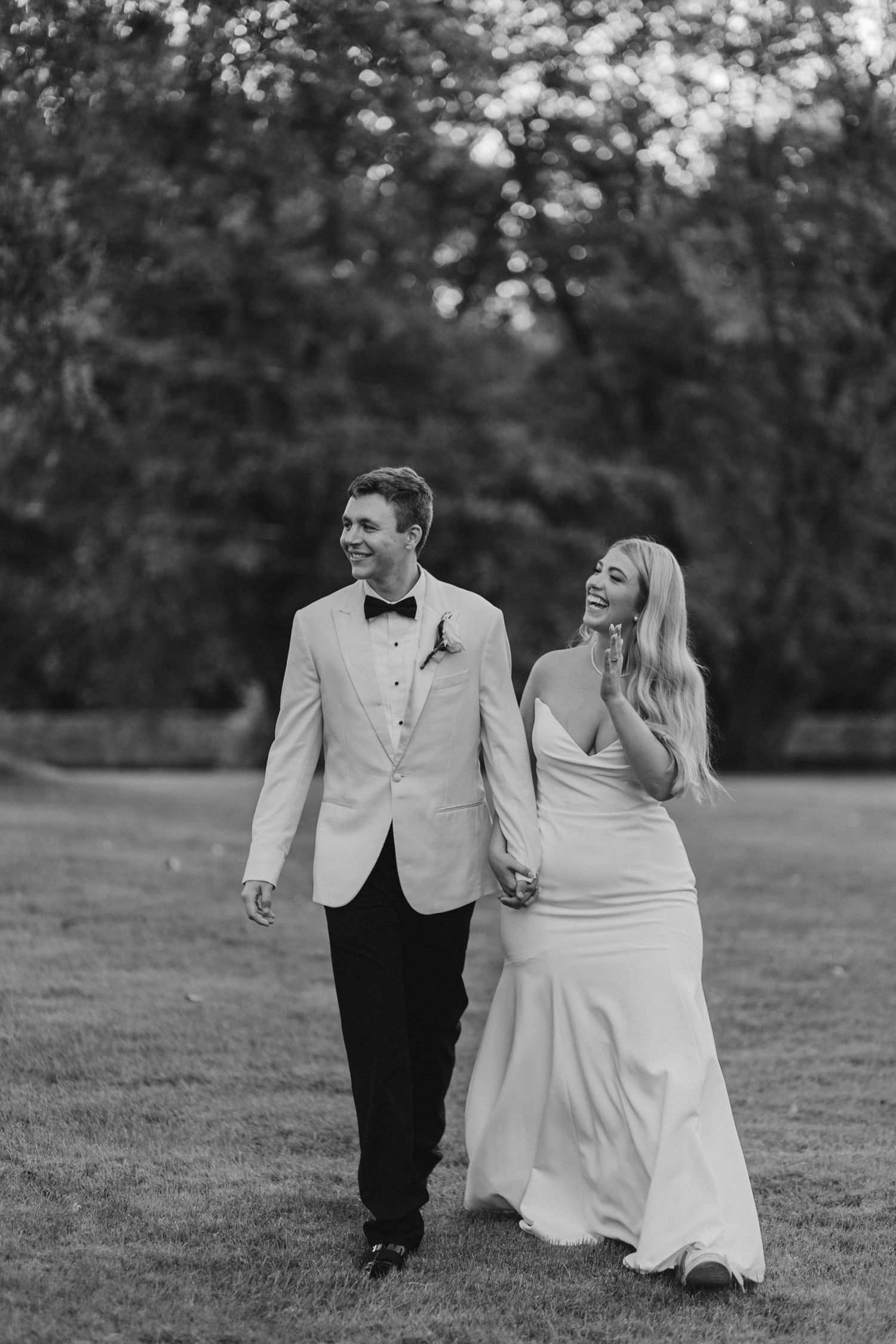 Black and white photo of smiling bride and groom walking hand in hand across a grassy field at dusk.