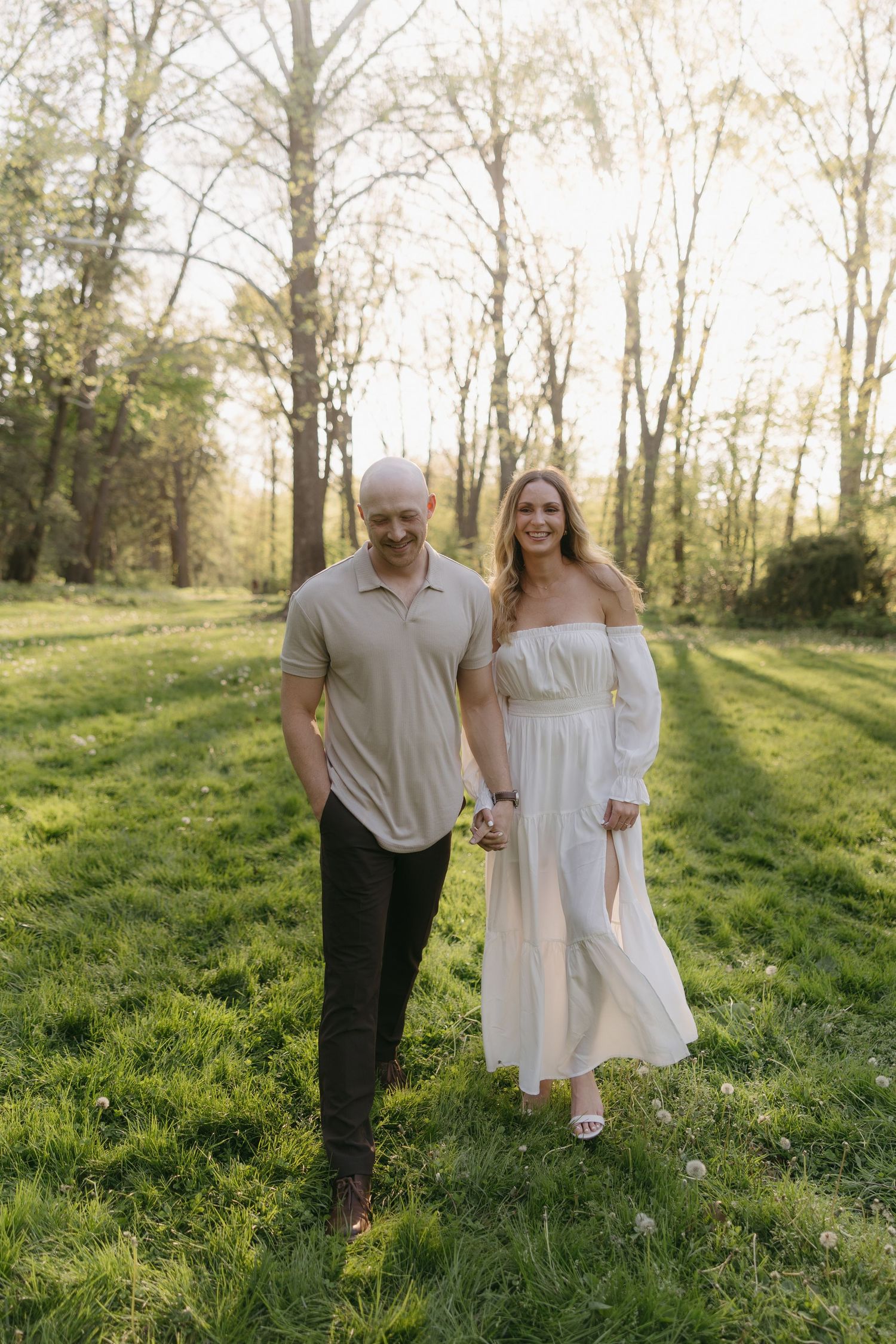 Smiling couple holding hands and walking through a lush green park at golden hour, woman in white dress.
