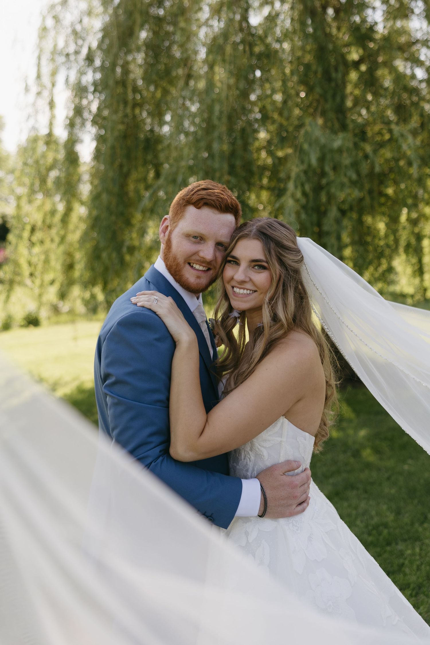 A smiling bride and groom embrace outdoors near a willow tree, her veil flowing in the breeze on their wedding day.