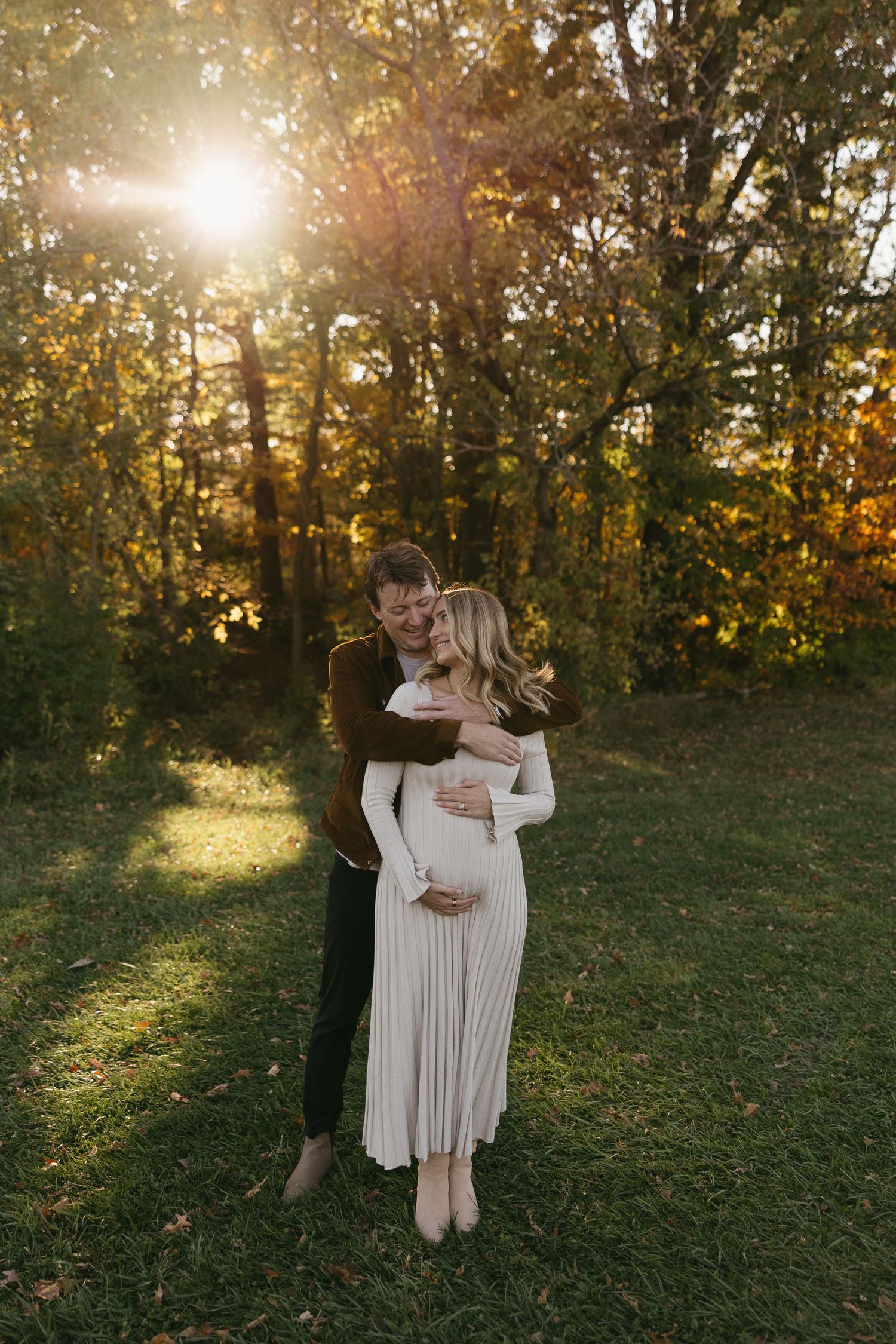Couple embracing outdoors during golden hour, pregnant woman in white dress, man in dark outfit, autumn trees background.