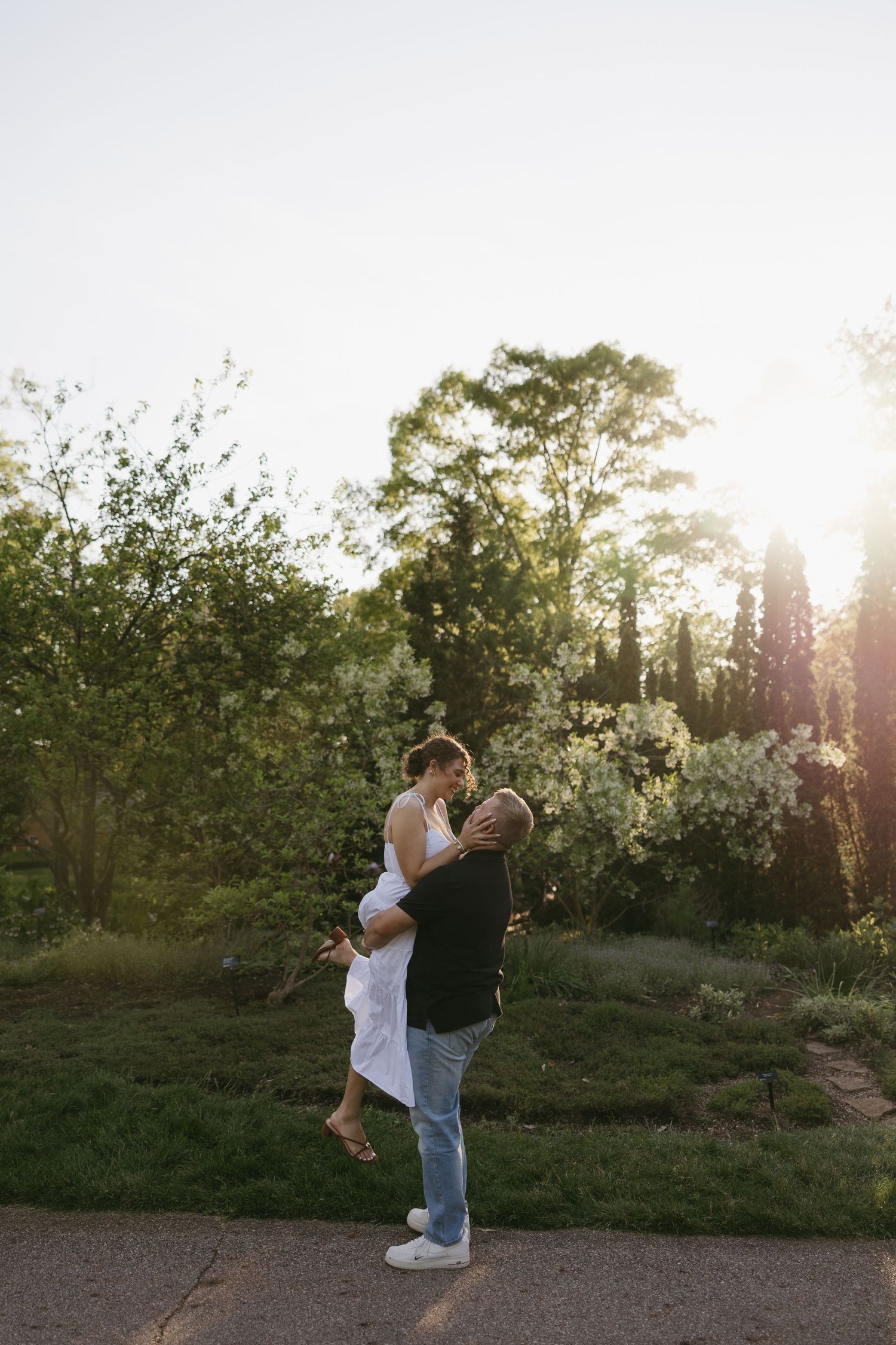 Couple embracing at sunset in a lush green garden, man lifting woman in white dress, golden hour light filtering through trees.