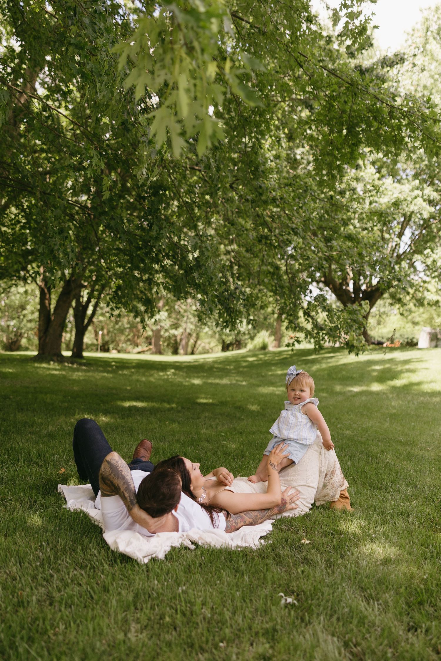 Family relaxes on white blanket in lush green park, parents lying down while baby girl sits up smiling.
